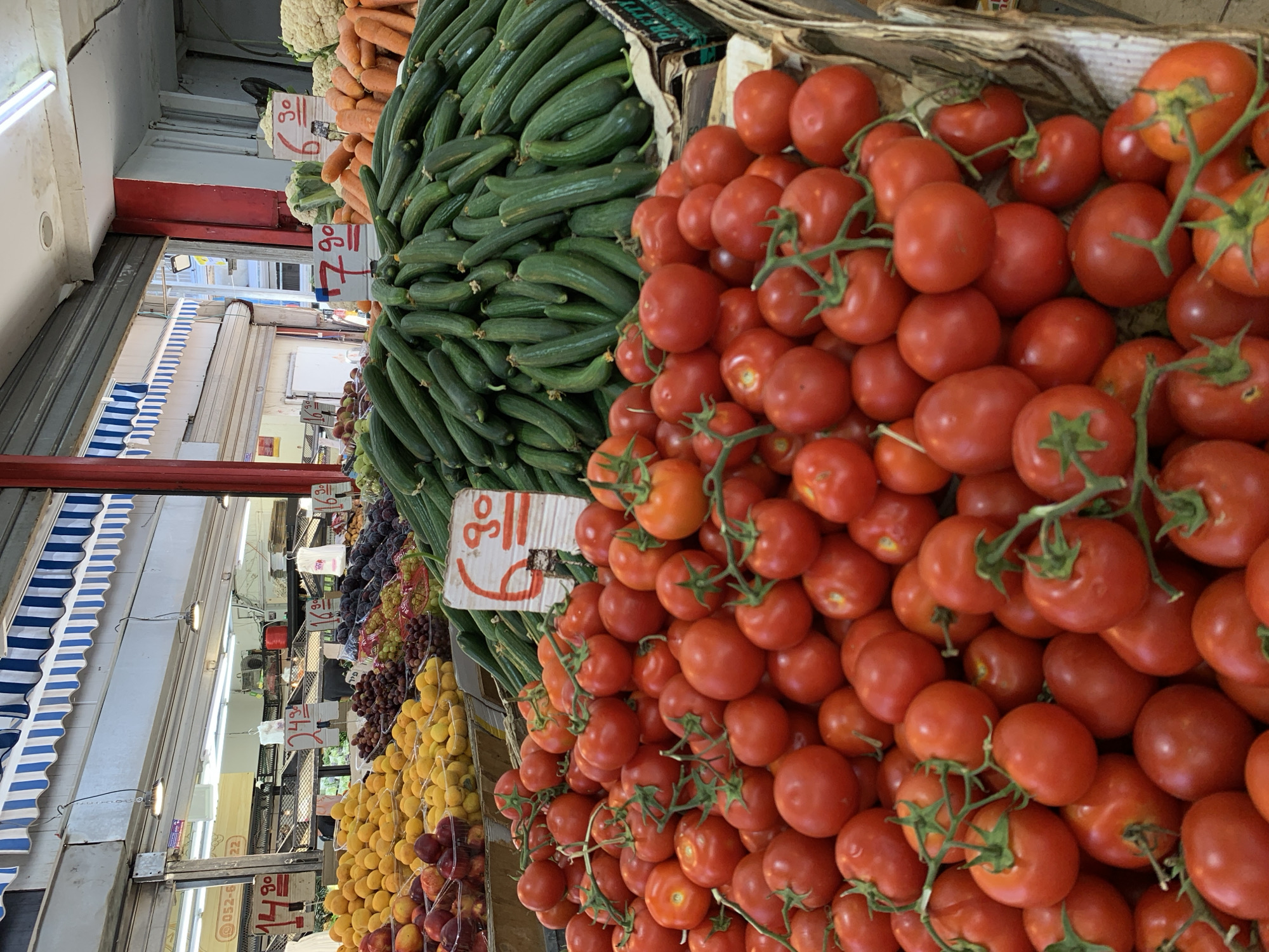 This is a street-level photograph taken at a produce market in Tel Aviv, Israel, likely in the Shuk HaCarmel (Carmel Market). The foreground is dominated by a large pile of ripe, red tomatoes. Behind the tomatoes, a substantial mound of green cucumbers is visible. Further back, there are displays of other fruits and vegetables, including peaches, plums, grapes, carrots, and cauliflower, suggesting a vibrant market scene. Striped awnings are visible in the background, typical of market stalls. Several price signs are placed throughout the market, indicating individual prices for the produce. The lighting suggests it is daytime.