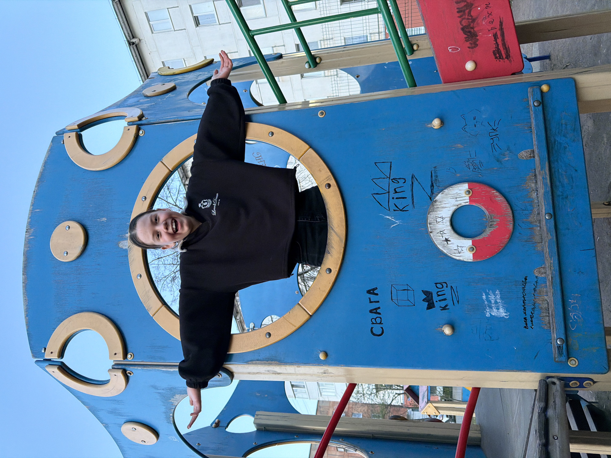 A young person is pictured on a blue and gold playground structure. The structure has circular openings, and the individual is positioned within one of these openings, extending their arms outwards. The background includes buildings, suggesting an urban or suburban setting. Graffiti is visible on the playground structure. The specific location is not discernible from the image.