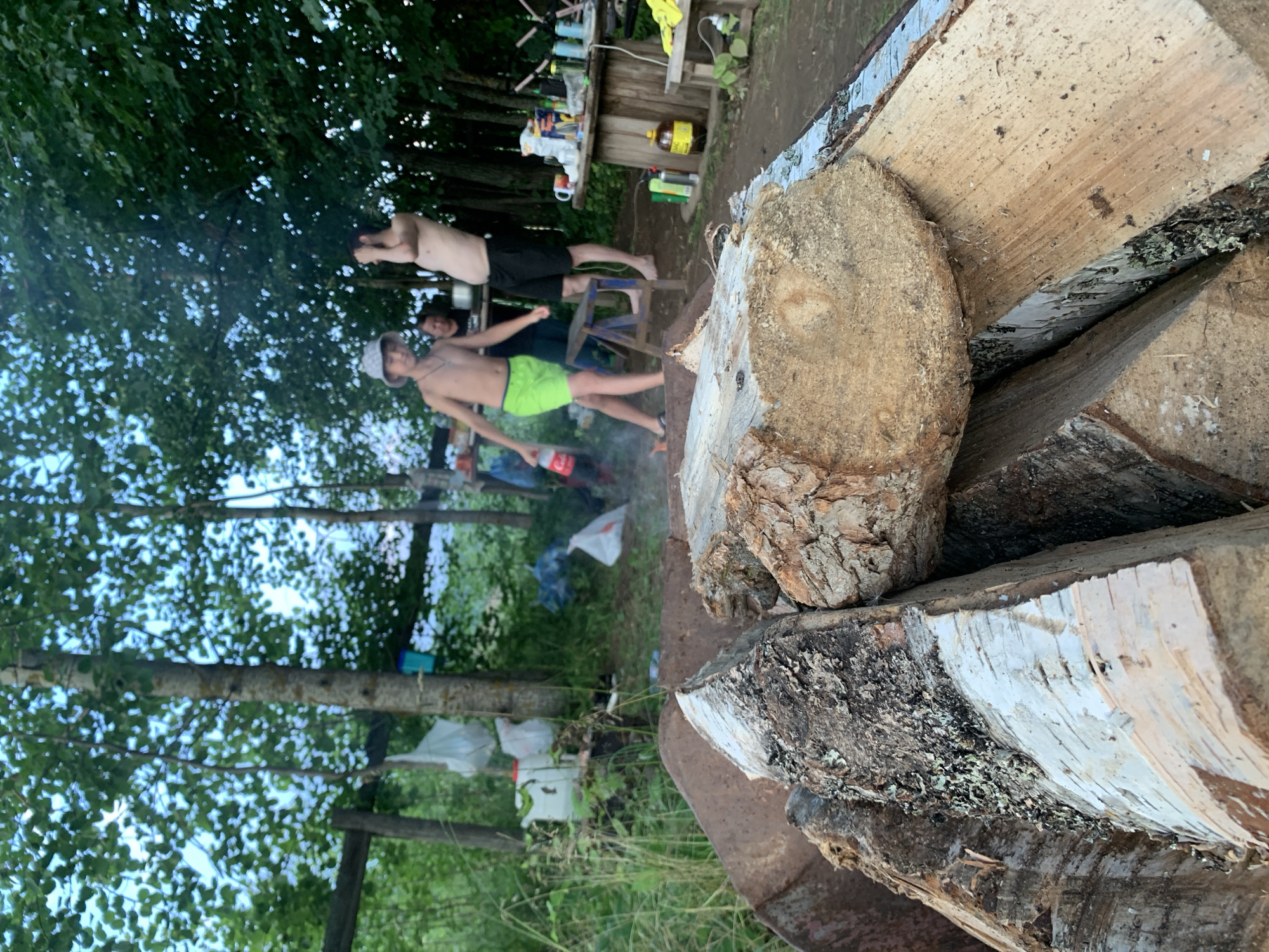 The image depicts an outdoor scene, possibly a campsite, with a foreground focus on a pile of chopped firewood inside a rusty metal basin. In the background, several people are visible. Two young males are standing, one wearing a hat and green shorts, holding a bottle of soda, and the other wearing black shorts. A third person, an adult female, is standing between them. The setting appears to be a wooded area with trees and foliage. Various items, including bags, containers, and a wooden table with supplies, are scattered around the background. The ground is dirt. The lighting suggests it is daytime, likely late afternoon or early evening. The presence of firewood implies preparation for a fire. There is no specific indication of the city of Zasechnyi, Russia, within the image itself.