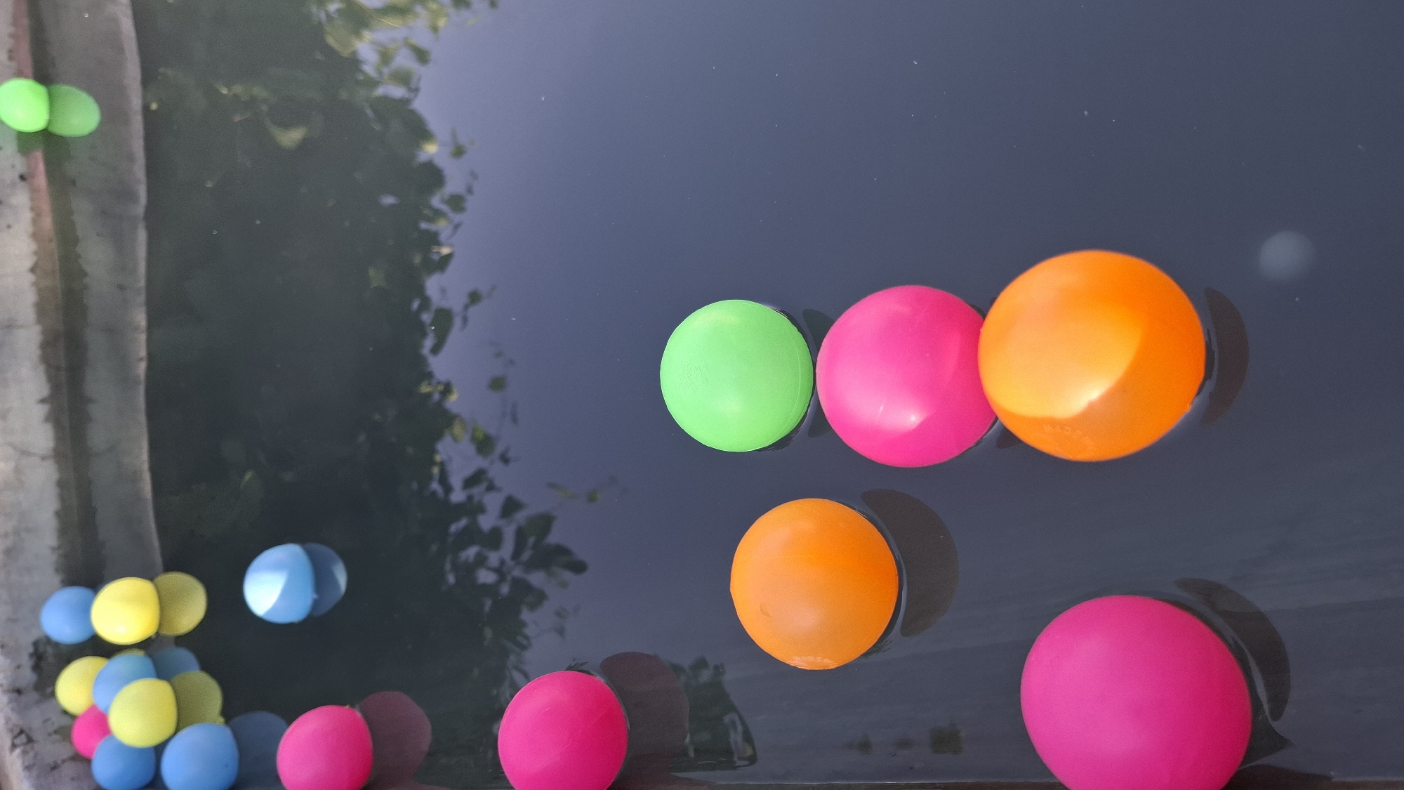 A close-up, vertical, high-angle shot shows several brightly colored plastic balls floating in dark water within a concrete pool. The balls are of various colors, including pink, yellow, blue, orange, and green. Some of the balls are clustered together near the edge of the pool, while others are more spread out. The water's surface is dark and reflective, showing faint reflections of foliage from above. The concrete edge of the pool is visible at the top of the frame. The scene appears to be outdoors. The specific location in the city of Шұбарсу, Kazakhstan is not discernible from the image itself.