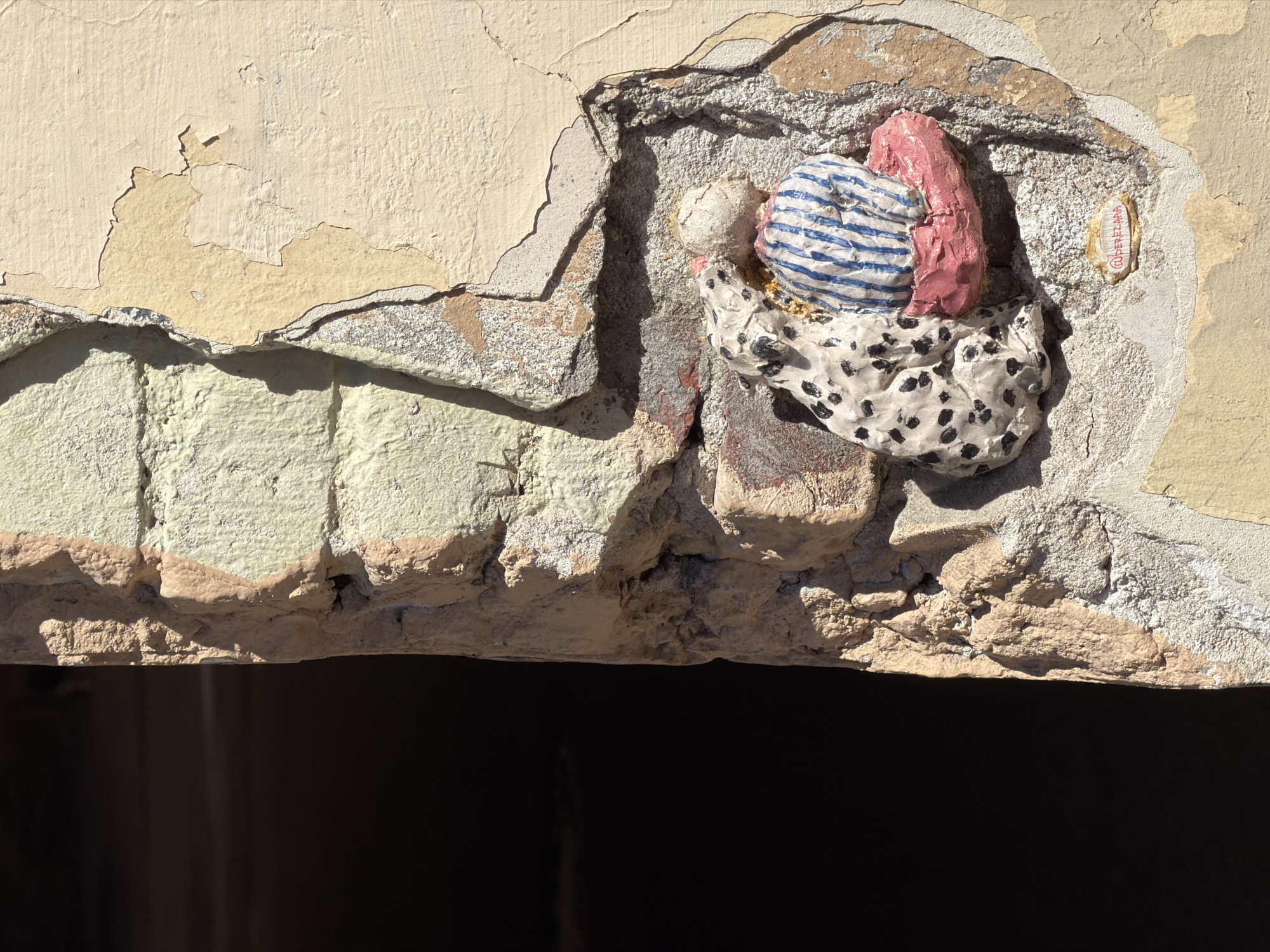 An exterior wall in Saint Petersburg, Russia, displays a sculptural relief embedded within a weathered recess. The wall exhibits multiple layers of light yellow-beige paint peeling away, revealing rougher, light tan plaster and underlying reddish-brown brickwork. The surface is textured and cracked.
The central sculpture, likely ceramic or clay, features two figures: a white, black-spotted dog (resembling a Dalmatian) on the left, shown in profile, appearing to lick or nuzzle the head of a second figure. The second figure, positioned on the right, is a rounded, anthropomorphic form with a light-colored, possibly bald, head and a torso adorned with vertical blue and white stripes, resting on a pink, cushion-like base. The sculpture is situated within a hollowed section of the wall where the plaster and brick are significantly exposed.
Below the sculpture, a small, oval-shaped tag is embedded in the wall, bearing partially legible red text that appears to read "@DEREZAKA."
Strong, direct sunlight illuminates the scene from the upper right, casting prominent shadows, particularly from the sculpture and the irregular textures of the wall. The left side of the image is dominated by a deep, undifferentiated black area, suggesting an unlit opening or architectural void adjacent to the wall.
