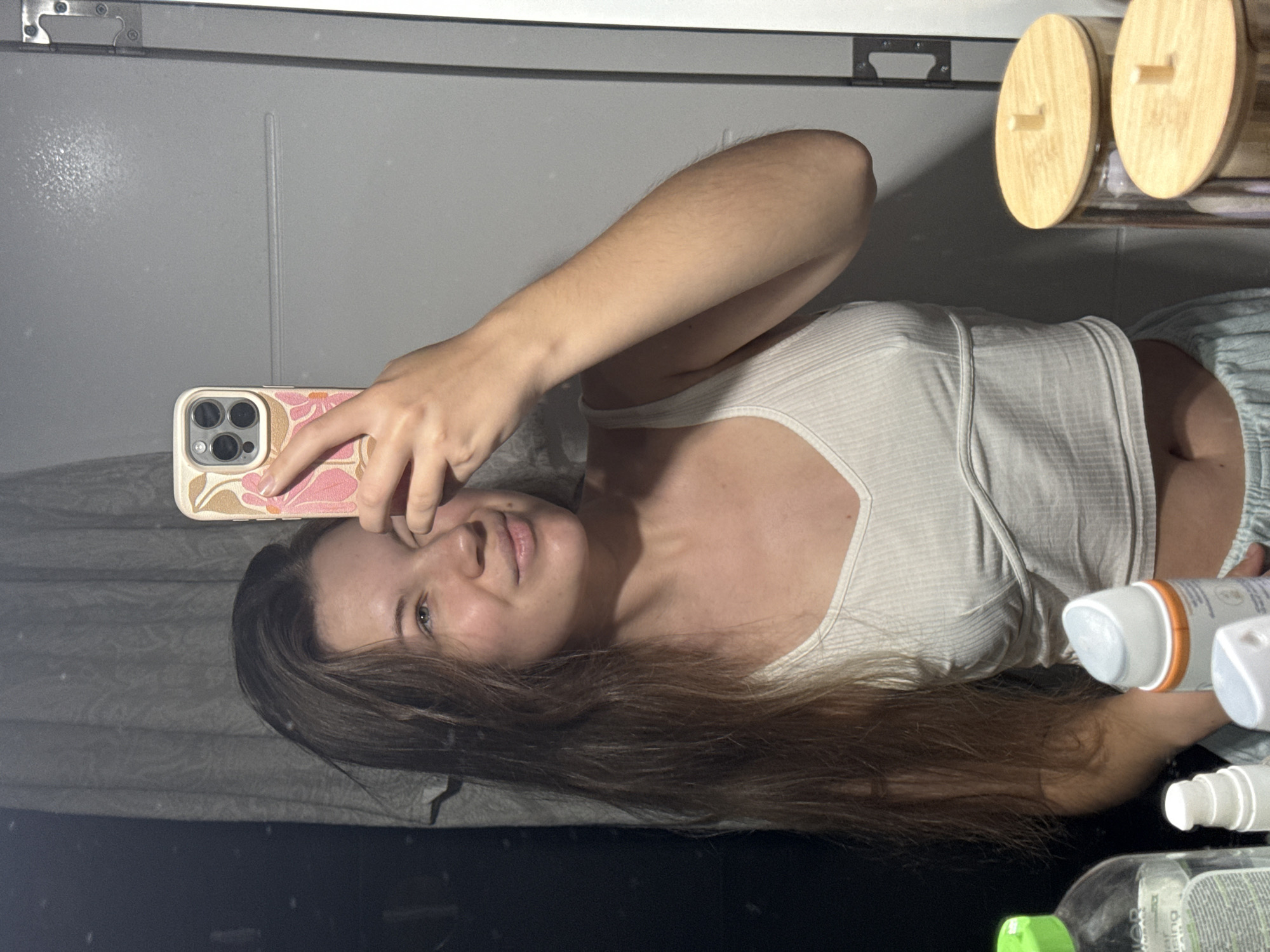 A young woman is taking a selfie in a mirror. She is wearing a white tank top and light blue shorts. Her hair is long and brown. She is holding a phone with a floral case in her right hand. On the counter near the mirror, there are storage containers with wooden lids and various bottles. The background shows a white wall. The location is not discernible from the image.