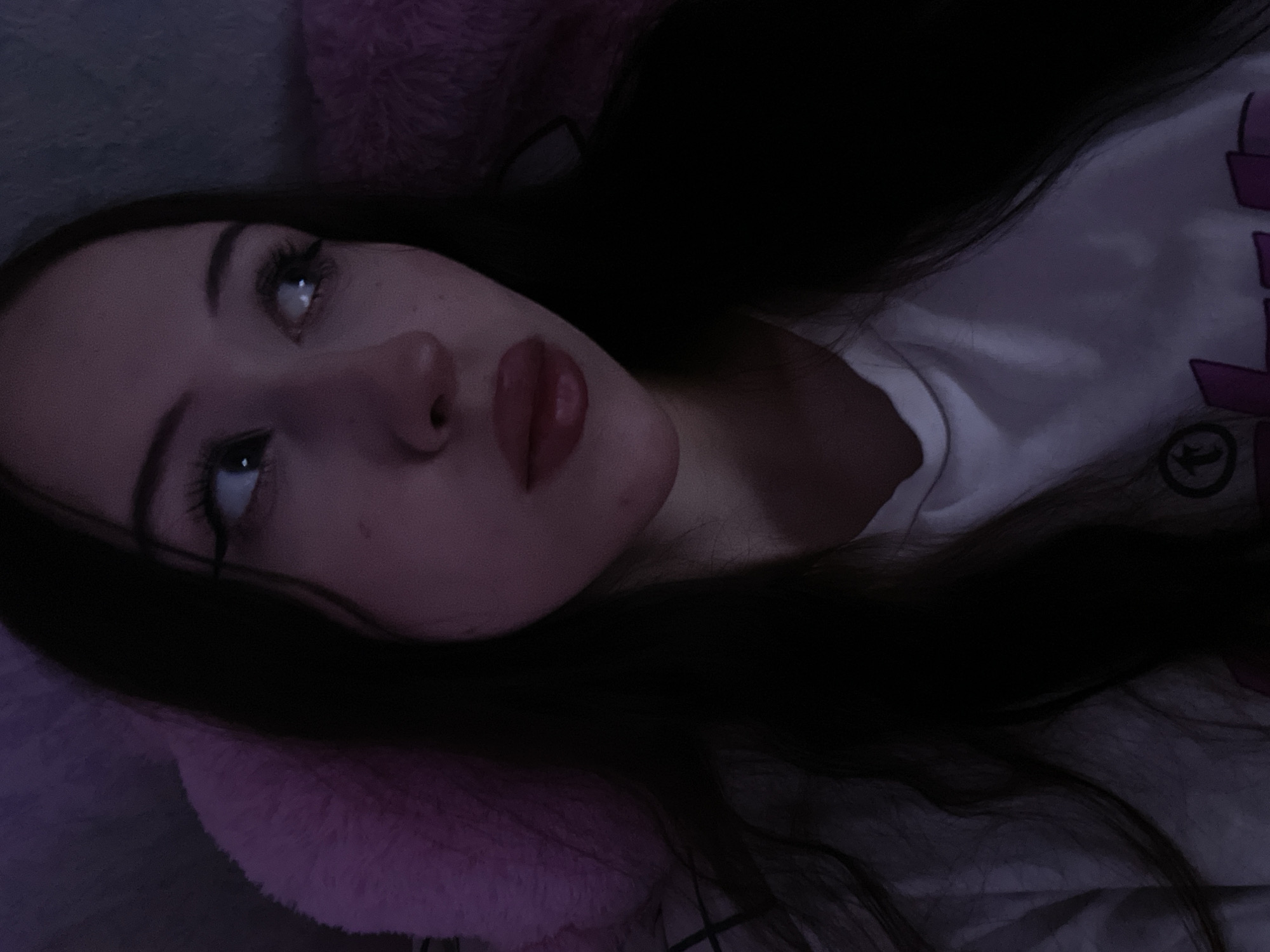 An undated, close-up selfie shows a young woman with long, dark hair, looking to her right. She is wearing a white t-shirt. The lighting is dim and purple-toned, suggesting an indoor setting, possibly at night. There is a fluffy pink object partially visible behind her head, which could be a pillow or part of a blanket. The image is cropped to focus on her face and upper torso. The context of Votkinsk, Russia is not evident in the visual information provided.