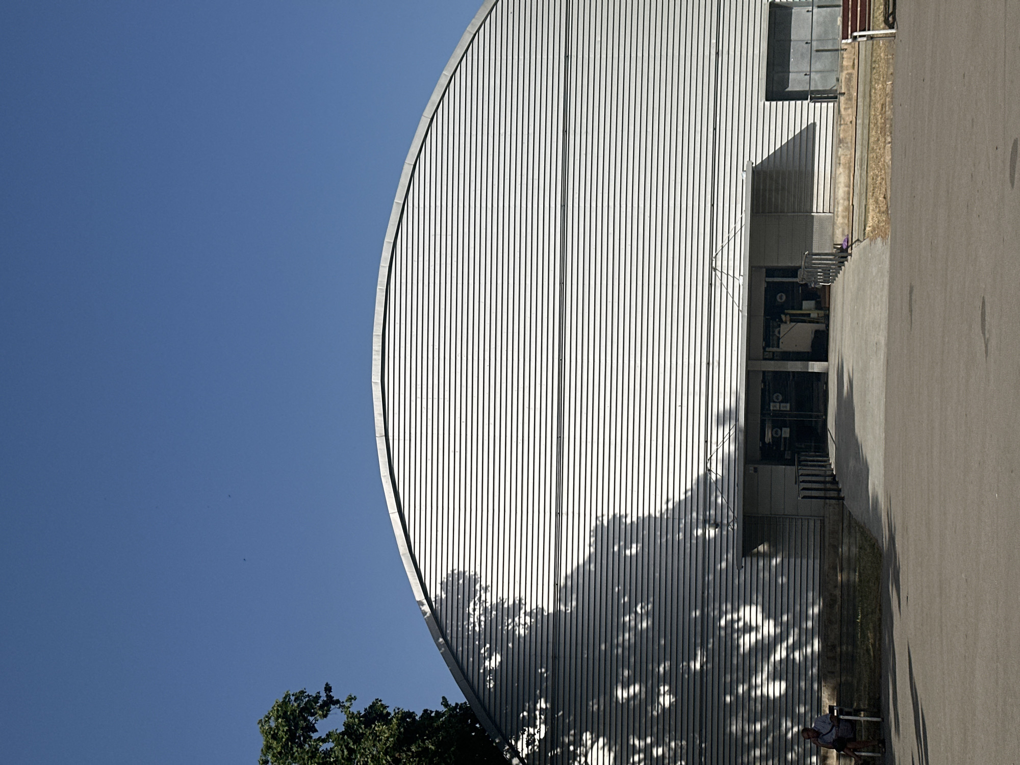 The image shows the exterior of a modern building with a curved, ribbed metal facade under a clear blue sky. The building has a large, arched entrance with glass doors and a ramp leading up to it. To the left of the entrance, a man is sitting on a bench, casting a long shadow from a nearby tree onto the building. The ground in front of the building is paved. The scene appears to be in an outdoor public space, possibly a park or plaza. The architecture suggests a contemporary design, and the presence of a tree and a bench indicates a space designed for public use.