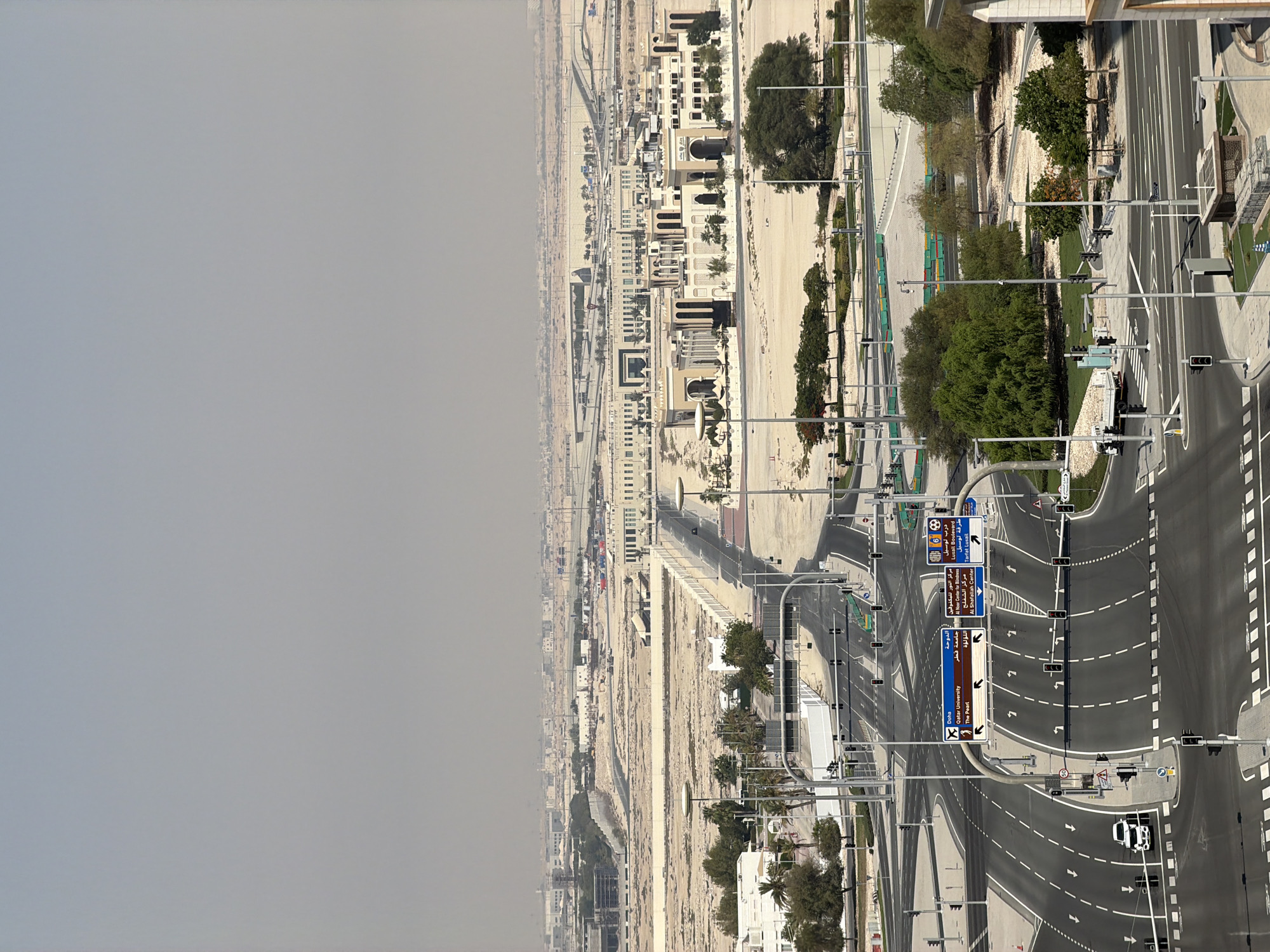 An aerial, eye-level, wide shot captures a modern cityscape under a hazy, light gray sky. In the foreground, a multi-lane intersection with a pedestrian crossing dominates the view. Traffic lights and signs, including one indicating directions to "Doha," "Qatar University," and "The Pearl," are clearly visible. Several cars, including a white SUV and a white truck, are present on the road.
Beyond the intersection, a long, wide road stretches into the midground, flanked by low-rise buildings with light-colored facades and arched windows, reminiscent of traditional Middle Eastern architecture. Palm trees and other sparse vegetation dot the landscape. The midground also features construction sites with barriers and machinery, suggesting ongoing development.
In the background, a sprawling expanse of beige, sandy terrain is dotted with more low-rise buildings and industrial structures. Elevated highways and overpasses can be seen in the distance, disappearing into the hazy horizon. The overall impression is one of a developing urban environment in a hot, arid climate.