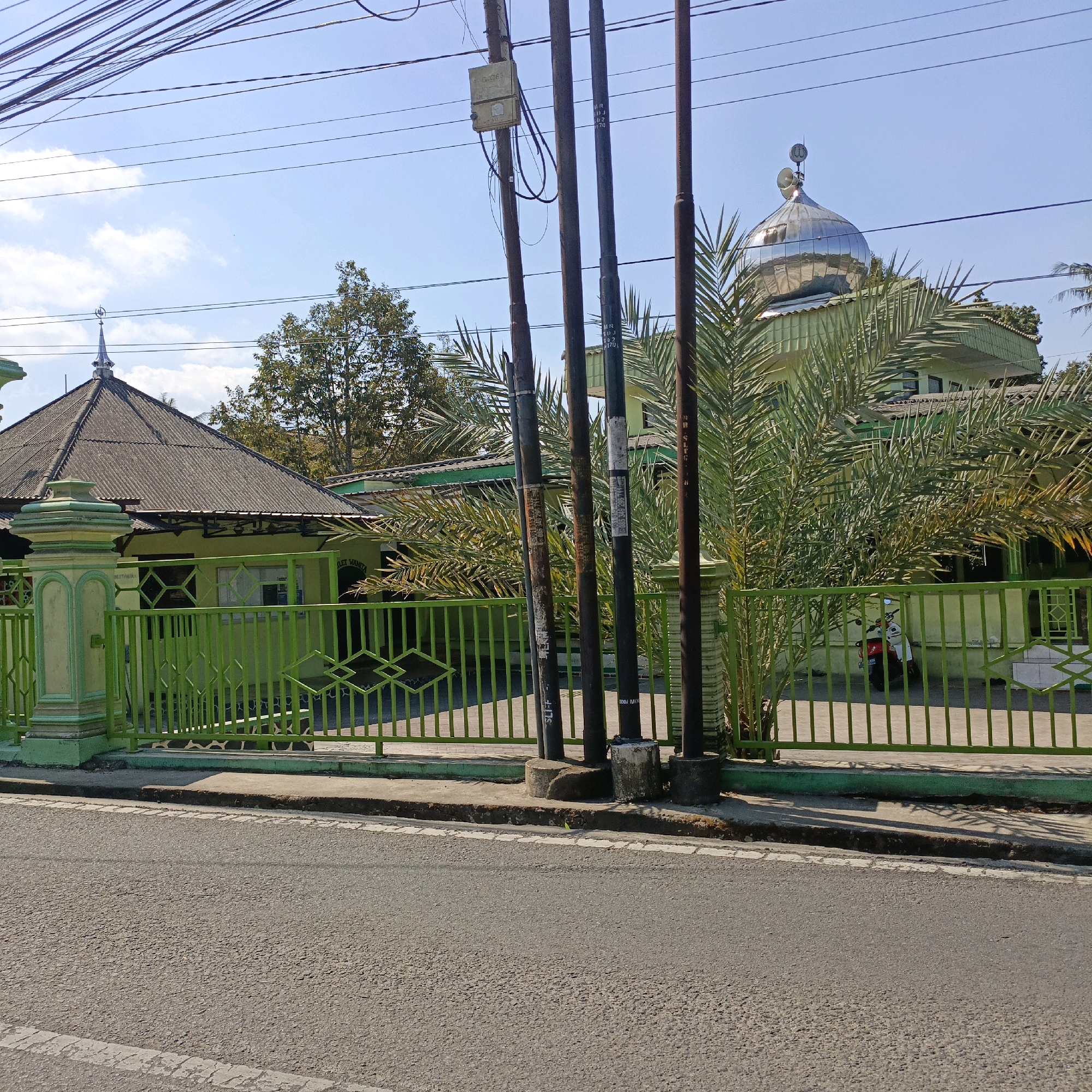 This image depicts the facade of a mosque in Salatiga, Indonesia, viewed from across a paved road. A bright green metal fence runs along the front of the mosque property, separating it from the road. Behind the fence, the mosque features a predominantly green building with a prominent silver dome. Palm trees with long, frond-like leaves are situated within the mosque grounds, partially obscuring the building. Several dark-colored utility poles, laden with a dense network of wires, stand in front of the fence and the mosque, running vertically through the frame. The sky is a clear blue with scattered white clouds. The foreground is dominated by the asphalt road, marked with white lines, and a concrete sidewalk. A red and white motorcycle is partially visible parked within the mosque grounds.