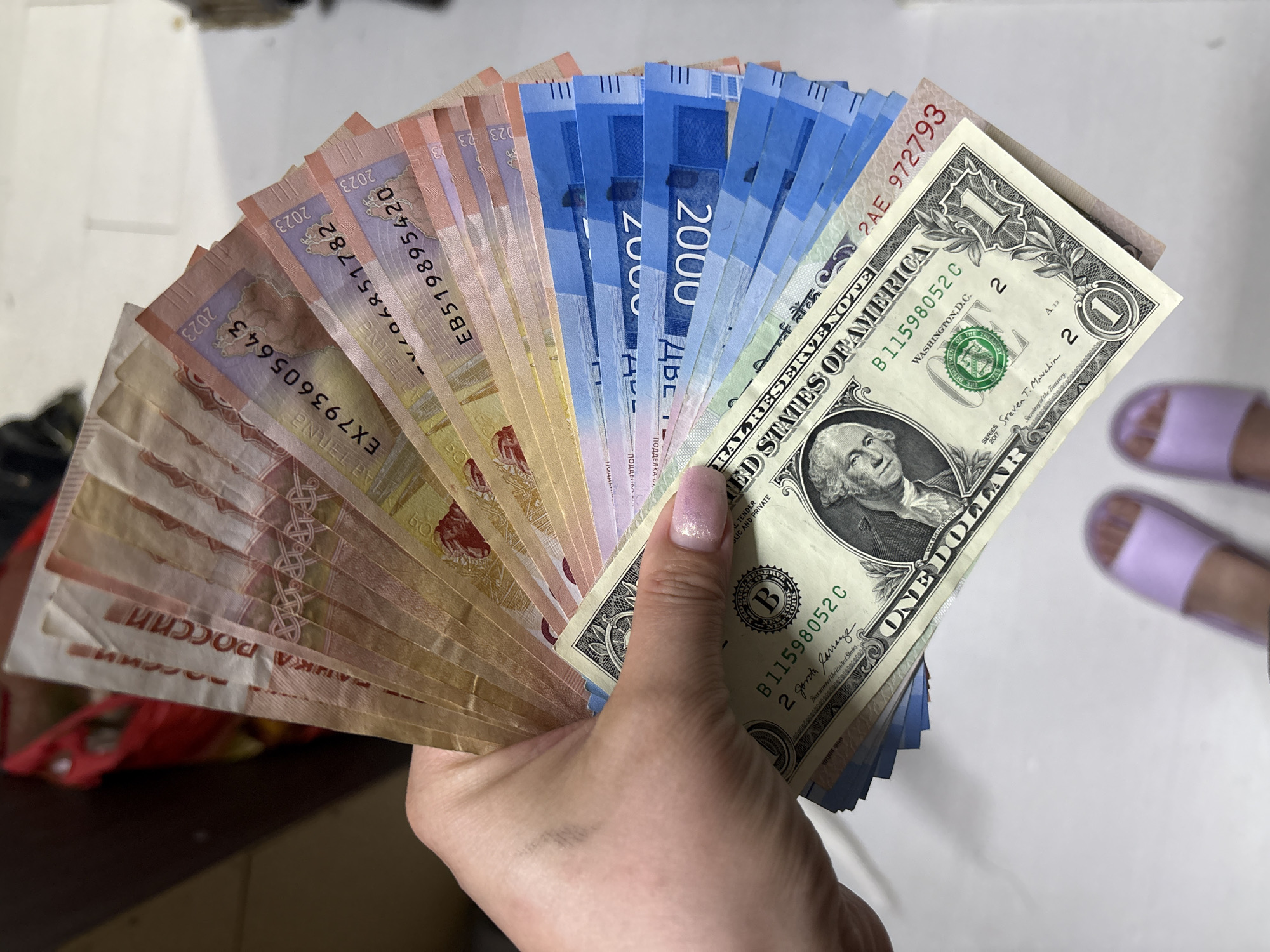A hand holds a fanned array of paper currency, including Russian Rubles of various denominations and a single US dollar bill. The rubles display the text "РОССИИ" and "2023". The dollar bill is a standard design. The hand holding the currency has a light-colored manicure. A pair of purple slippers is visible in the upper right corner of the image. The background is a light-colored surface. The currency includes blue 2000 notes, and pink and yellow 5000 notes. There is no explicit indication of the location Марковский, Russia, in the image.