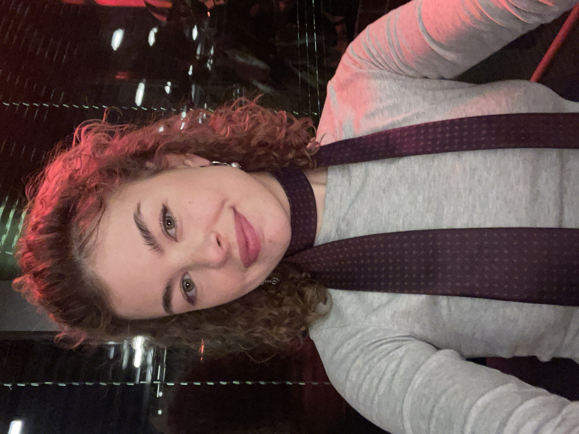 A woman is pictured indoors, likely in an entertainment venue, possibly in Tbilisi, Georgia. The environment is dark with blurred lights in the background, potentially a tunnel or decorative feature with mirrored walls. The woman has light skin, curly reddish-brown hair, and is wearing a grey long-sleeved shirt with a dark burgundy patterned tie or scarf around her neck. She is looking at the camera with a slight smile. The image orientation is rotated clockwise.