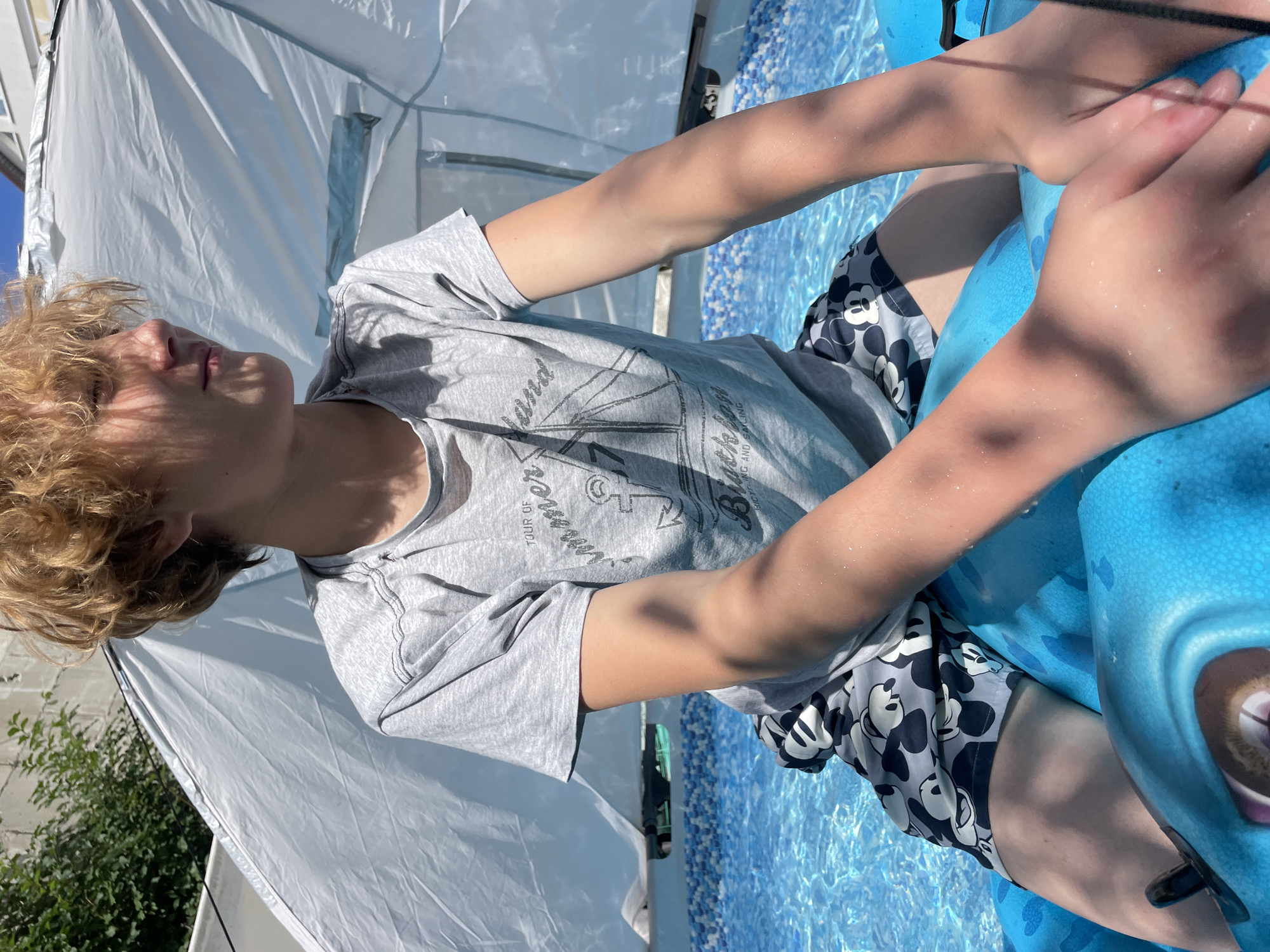 A young person with blond, messy hair is lying on their side in a bright blue inflatable pool filled with clear water. They are wearing a grey t-shirt with a graphic design of a sailboat and text that reads "TOUR OF SUMMER" and "BICYCLE" with a line underneath. They are also wearing grey swim trunks with a pattern of Mickey Mouse faces. The person's eyes are closed, and their head is tilted back, suggesting relaxation or sleep. Sunlight casts dappled shadows on their skin and clothes, indicating it's a sunny day. In the background, a large white tent or canopy provides shade. The setting appears to be an outdoor area, possibly a backyard, with greenery visible to the left and a blue tiled edge of the pool in the foreground. The overall mood is peaceful and summery.