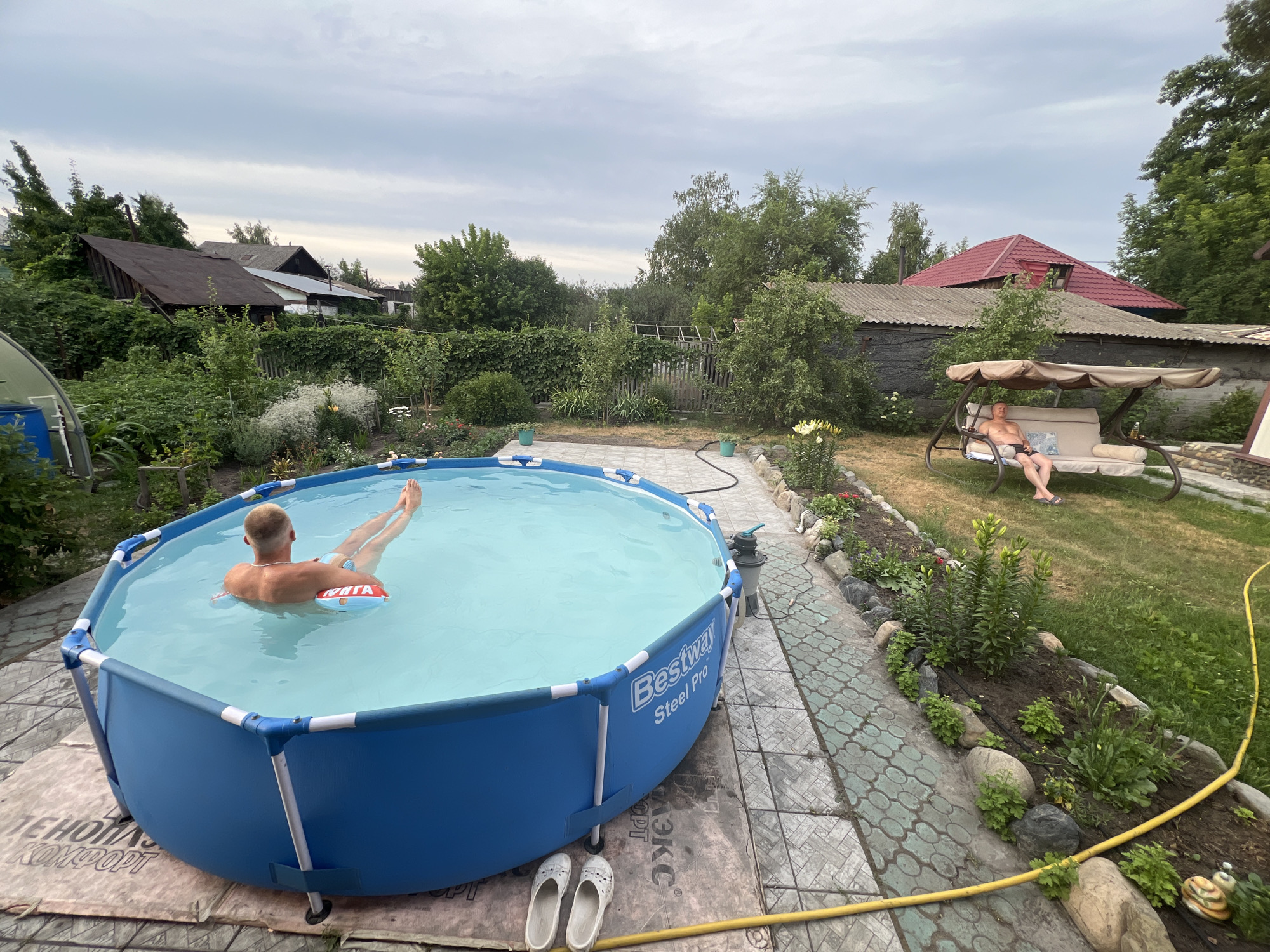 A man is relaxing in an inflatable pool in the yard of a house. He is partially submerged in the water, with his head and shoulders visible above the surface. He appears to be floating on a small inflatable ring. The pool is blue and round, with a white frame. It is set up on a paved area in a backyard with lush greenery and trees. In the background, there is a house with a red roof and a swing set with another person sitting on it. The sky is cloudy and overcast. The image is taken from a slightly elevated angle, looking down at the pool and the man.