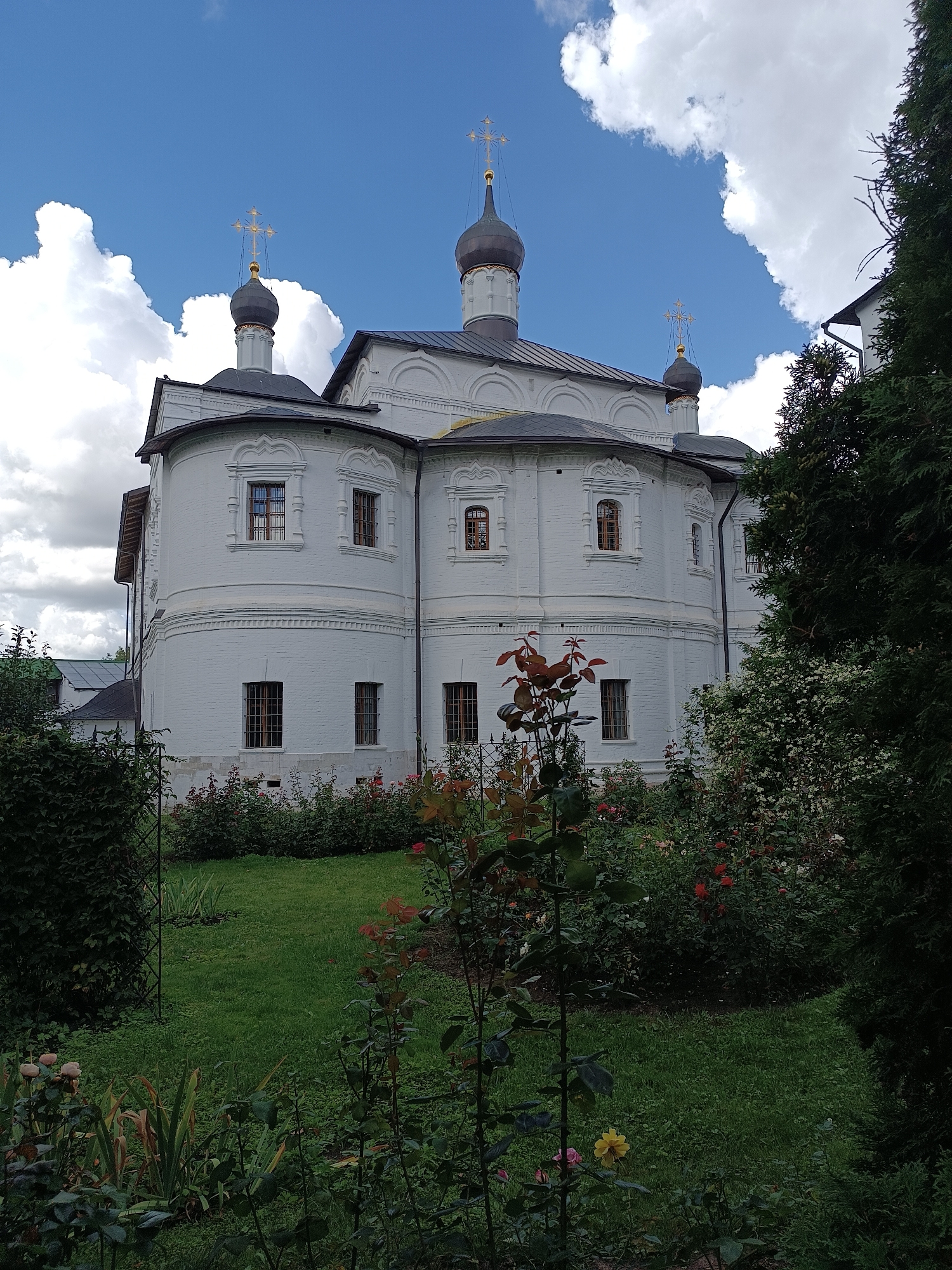 This is an eye-level, medium shot of the Trinity Lavra of Saint Sergius in Sergiyev Posad, Moscow Oblast, Russia. The exterior of the white brick church, with its dark domes topped with golden crosses, is visible against a blue sky with fluffy white clouds. The church has several windows with ornate frames and wrought-iron grates. In the foreground, a lush green lawn is surrounded by a variety of plants and flowers, including roses, with trees framing the right side of the image.