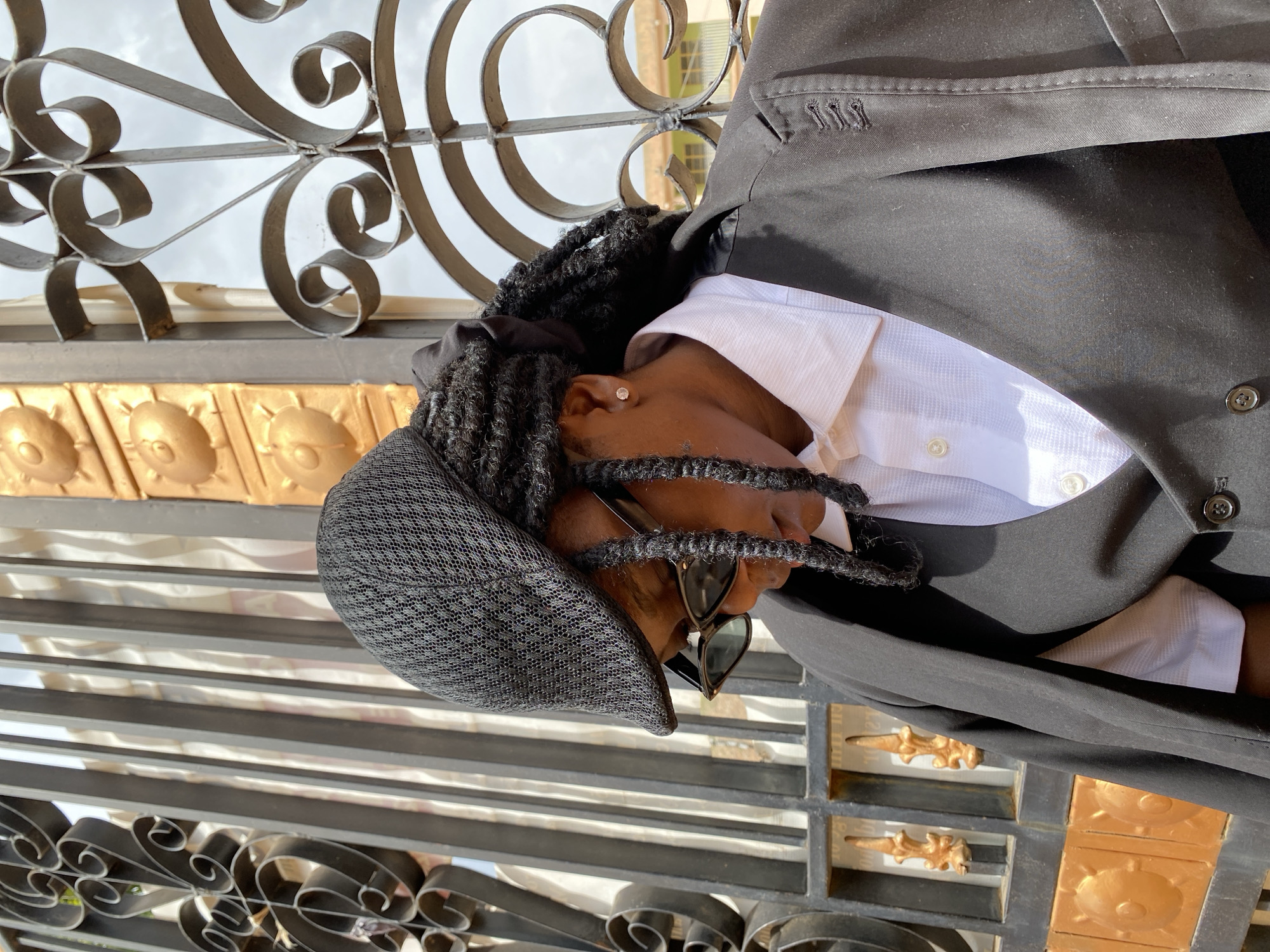 An individual stands in front of an ornate metal gate with gold accents. The person wears a dark-colored suit, a white collared shirt, a patterned cap, and sunglasses. Their hair is styled in braids. The gate is located in Gwong, Nigeria.