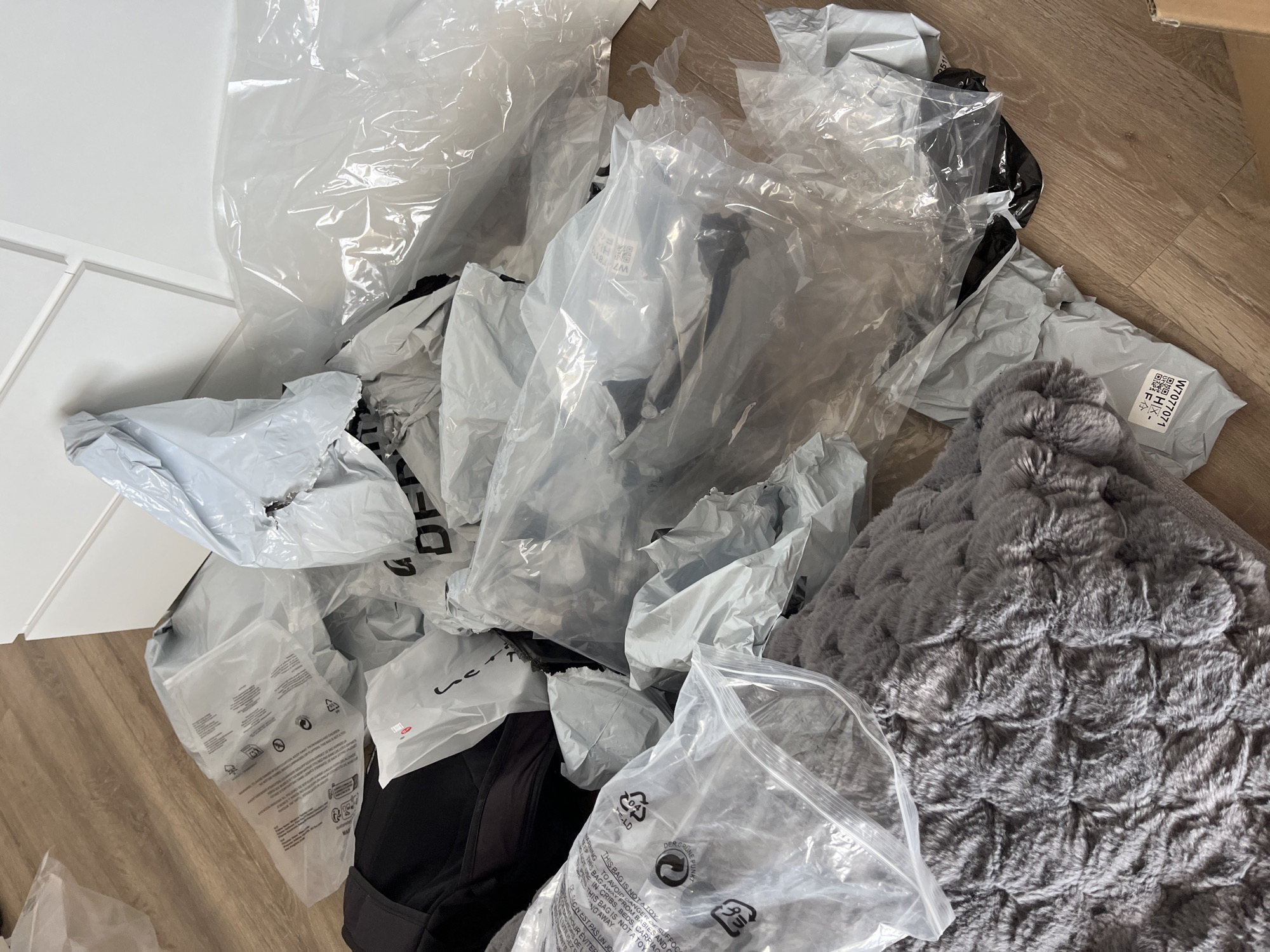 The image shows a collection of packaging materials and clothing items scattered on a light brown wooden floor. Predominantly, there are numerous crumpled and translucent plastic bags, some with printed text and recycling symbols. Beneath and partially obscured by the bags are sections of black clothing. To the right, a textured, gray faux fur item, possibly a blanket or cushion, is also visible. A partial view of white furniture is also visible in the image to the left. The scene does not show any specific details that would suggest it is in Zlatograd, Bulgaria. The details are general and could be situated in any similar domestic setting.