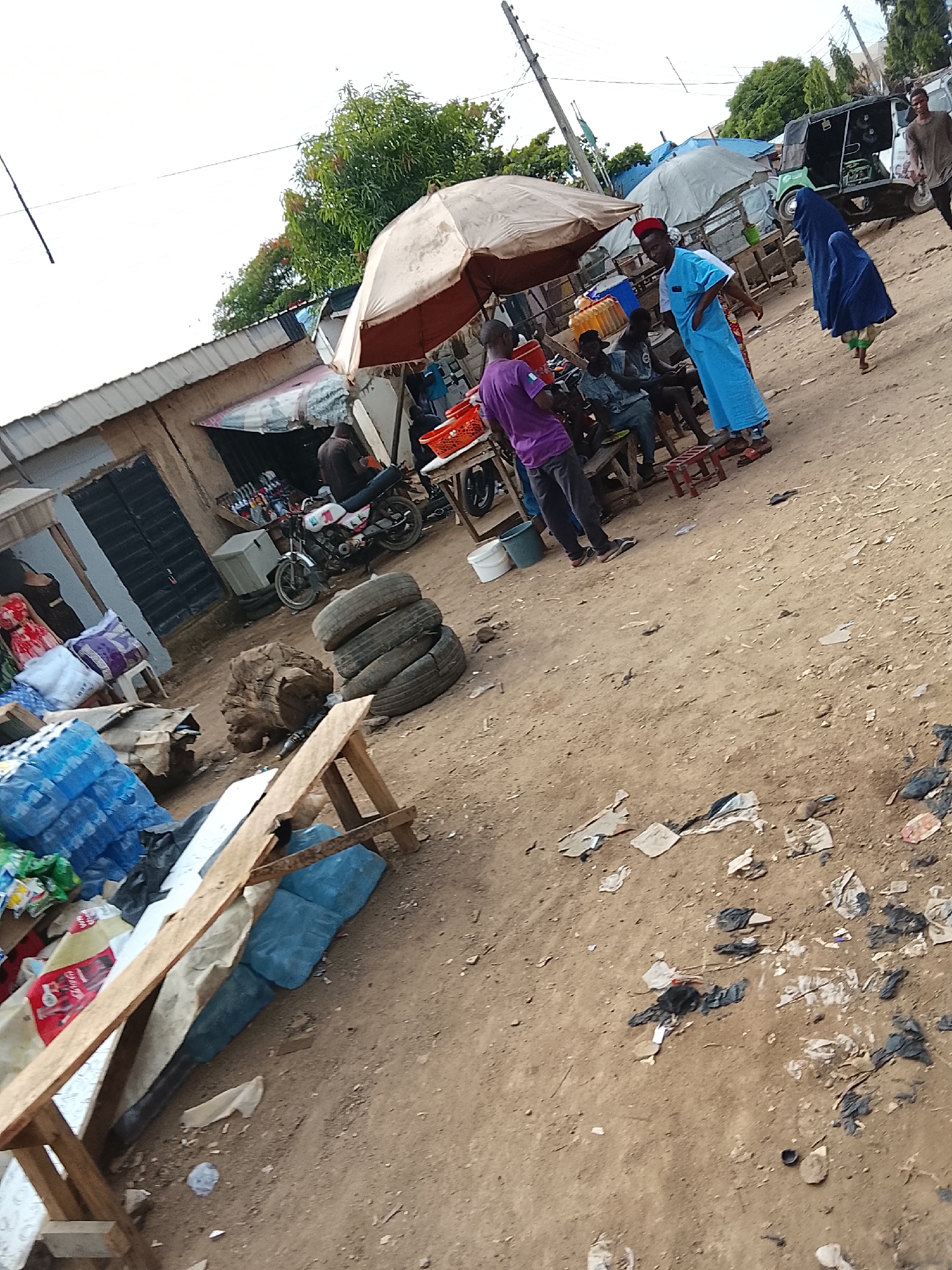 The image depicts a street scene, potentially a marketplace, in Abuja, Nigeria. A group of people are gathered under a large, light-colored umbrella, some sitting on benches and stools. A man in a bright blue robe and a red cap stands, possibly engaged in conversation. A woman wearing a dark blue hijab walks nearby.
A motorcycle is parked in front of a building. A stack of used tires and a large piece of wood are nearby. In the foreground, there are stacks of bottled water and a makeshift wooden bench or rack. Litter is scattered on the dirt ground.