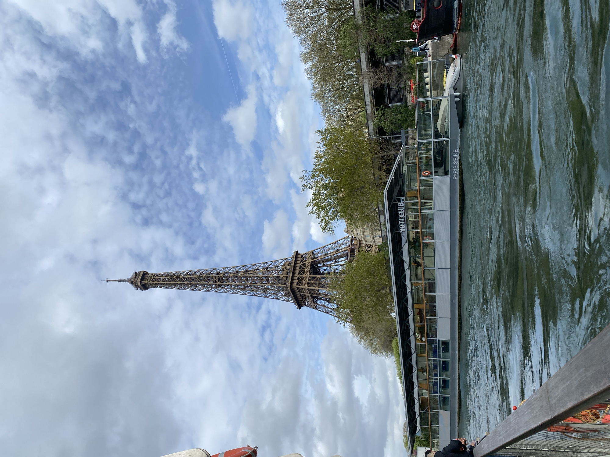 The image depicts the Eiffel Tower in Paris, France, viewed from a boat on the Seine River. The tower is positioned on the left, reaching into a partly cloudy sky. Adjacent to the river is a boat called "Paris Seine", used as a club. A walkway is visible in the foreground. The river itself has ripples and reflections. Trees line the riverbank in the background.
