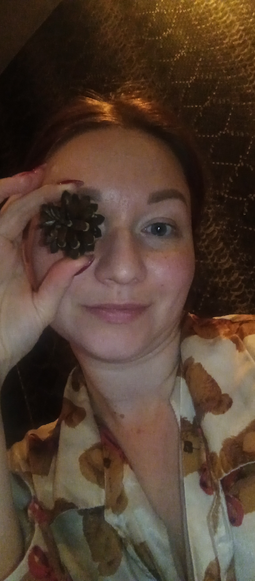 A woman is holding a pine cone up to her left eye. She has fair skin, red hair, and blue eyes. She is wearing a cream-colored pajama top with brown teddy bears printed on it. Her nails are painted red. The background is blurry, but it appears to be a dark brown, patterned surface. The location appears to be inside, but the specific city and location are unknown.