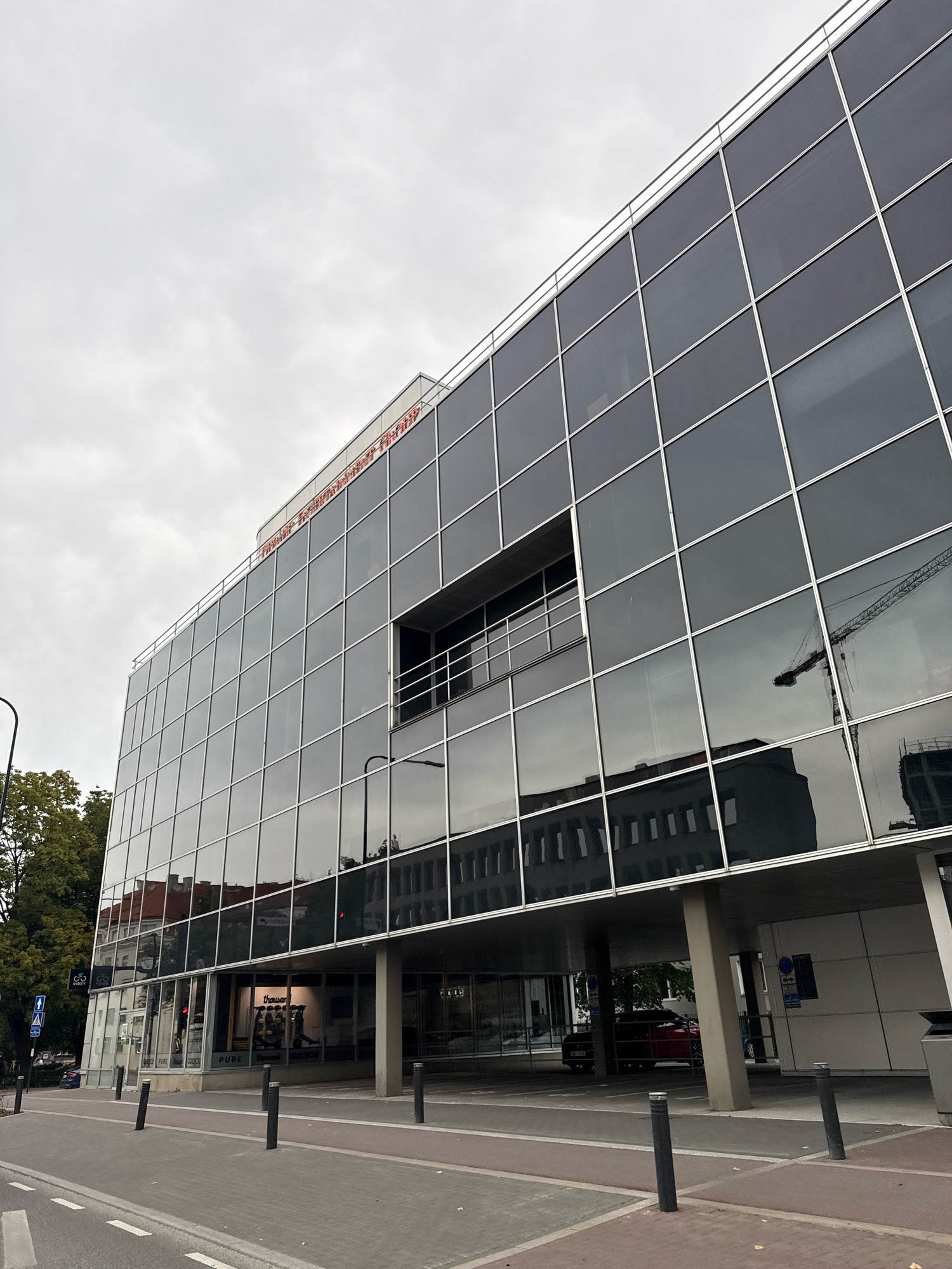 The image shows the exterior of a modern office building with a glass facade in Tallinn, Estonia. The building has a prominent red sign on its roof that reads "TALLINNA LINNAVALITSUS" which translates to "Tallinn City Government". The glass facade reflects the surrounding environment, including a construction crane and other buildings. Below the office levels, there are retail spaces with storefronts. A red car is parked under the overhang of the building. The street in front of the building has a pedestrian walkway and a road with lane markings. Trees and a street lamp are also visible. The sky is overcast.