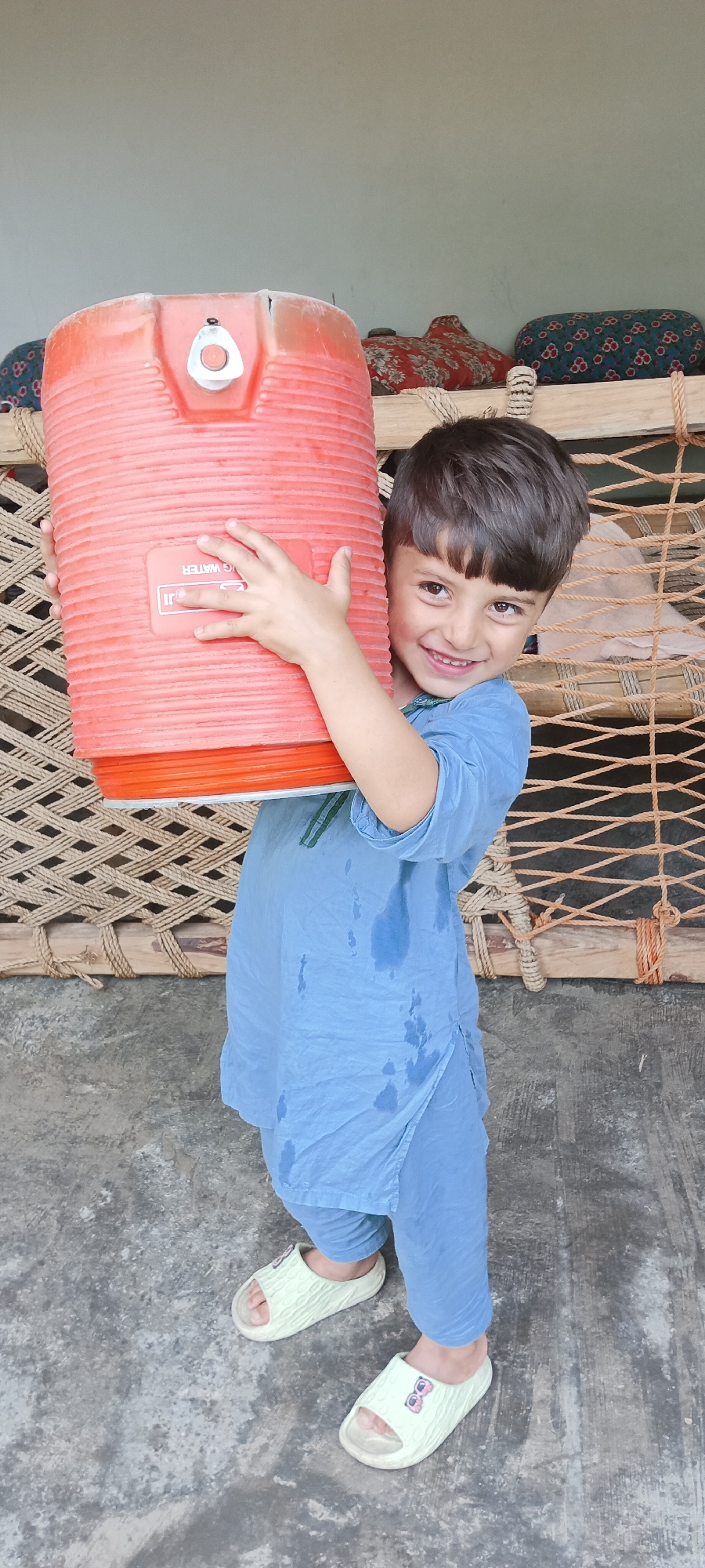 A young boy stands outside in Katlang Tehsil, Pakistan, holding a large, red water cooler. He wears a blue shalwar kameez, which is wet on the front, and green sandals. The background includes a woven charpai and colorful patterned cushions on a raised platform. The flooring is gray concrete.