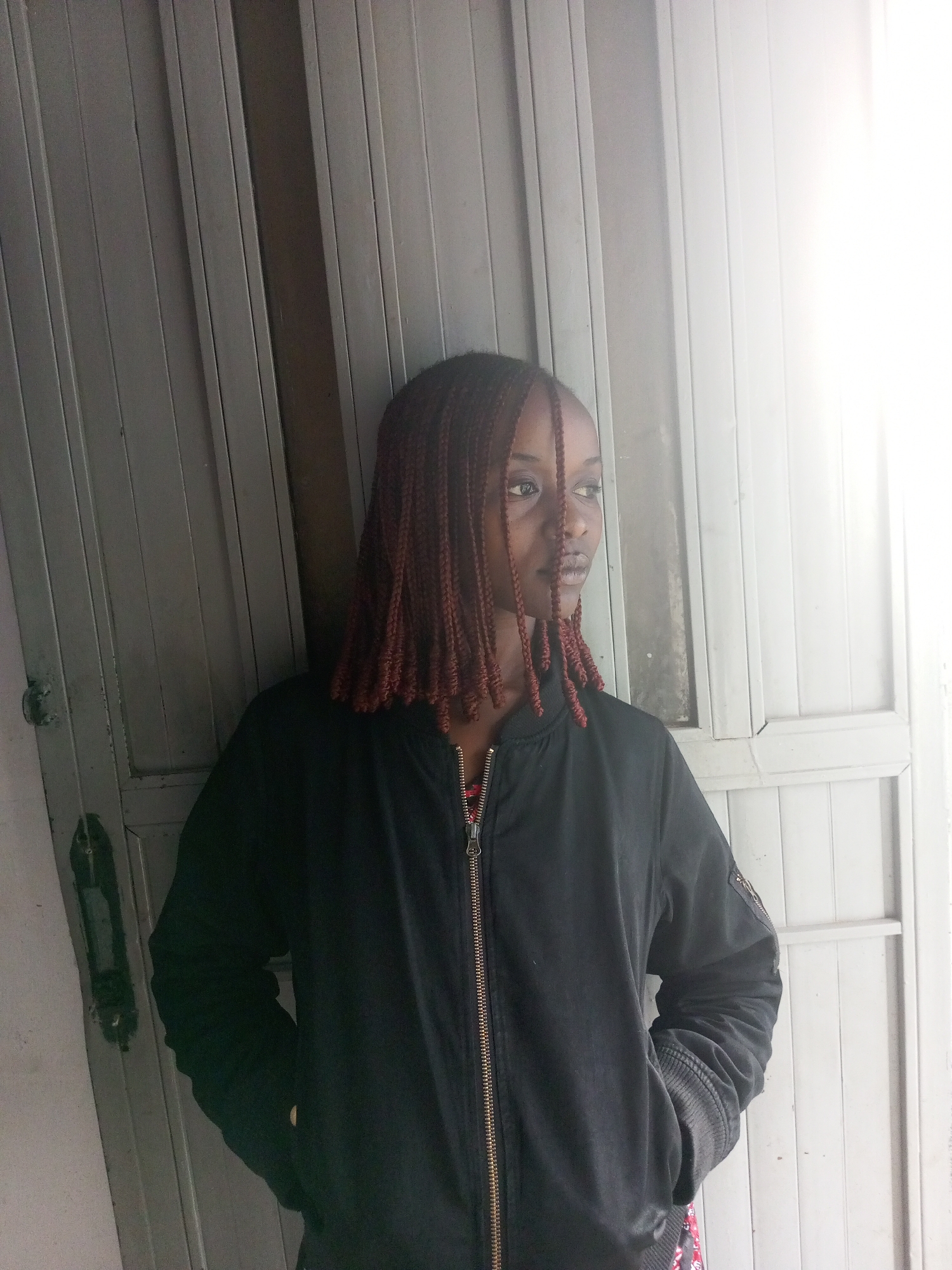 A medium shot shows a young woman with braided, dark red hair standing in front of a gray, weathered metal door. She is wearing a black bomber jacket with the zipper partially undone, revealing a hint of a red and white patterned shirt underneath. Her hands are tucked into the pockets of her jacket. The lighting on the right side of the image is bright and washed out, suggesting a light source such as a window or open doorway. The background is dominated by the vertical lines of the door. The overall scene appears to be an outdoor or semi-outdoor setting.