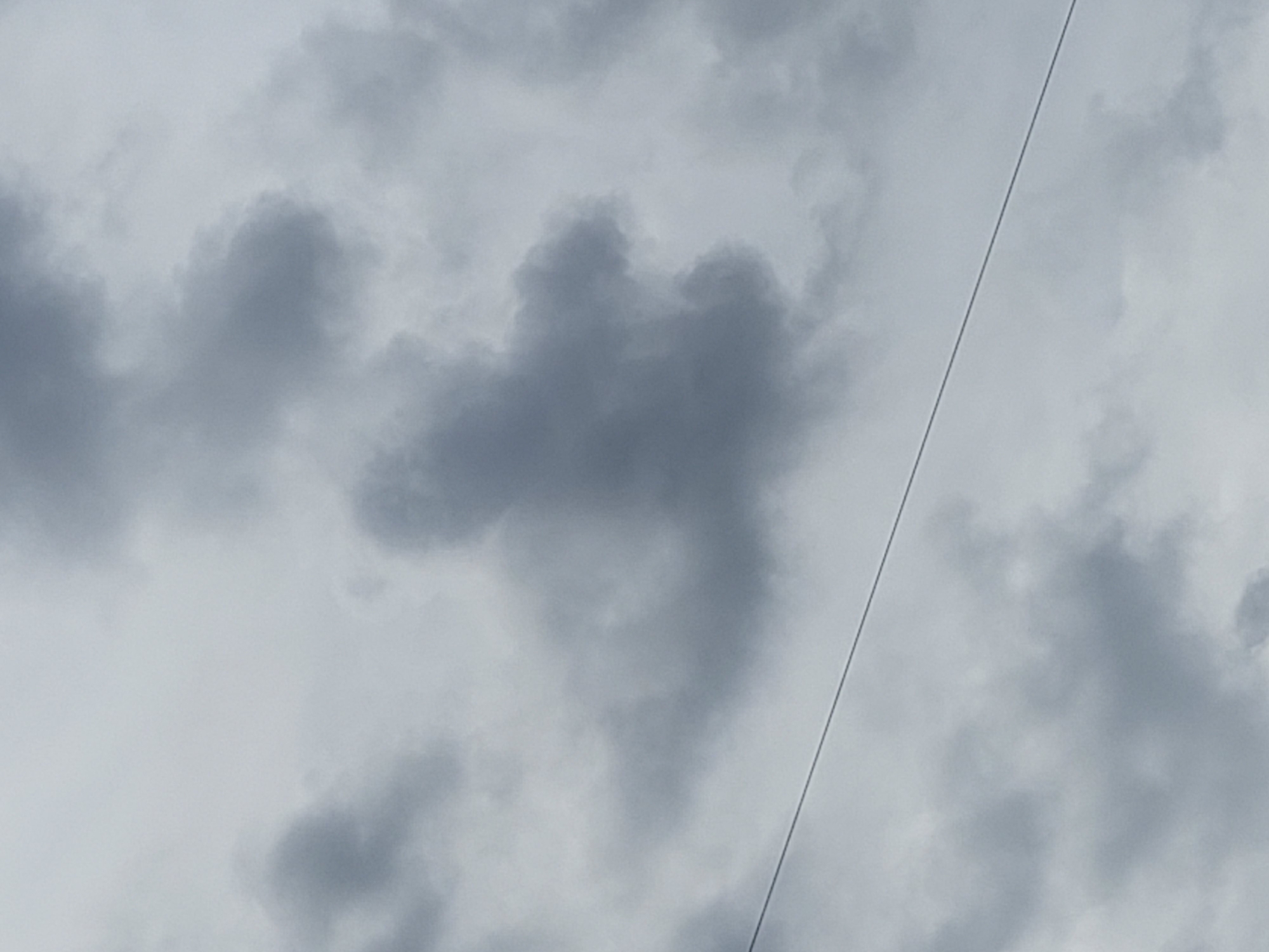 The image shows a cloudy sky with a wire extending diagonally across the frame from the bottom left to the top right. The clouds are a mix of dark and light gray, indicating overcast conditions. There are no people or visible structures in the frame, besides the wire. The setting is an outdoor environment, likely in the city of Unknown, Unknown, given the presence of a wire that could be part of an electrical or communication line. The overall mood is somber and neutral due to the dull weather.