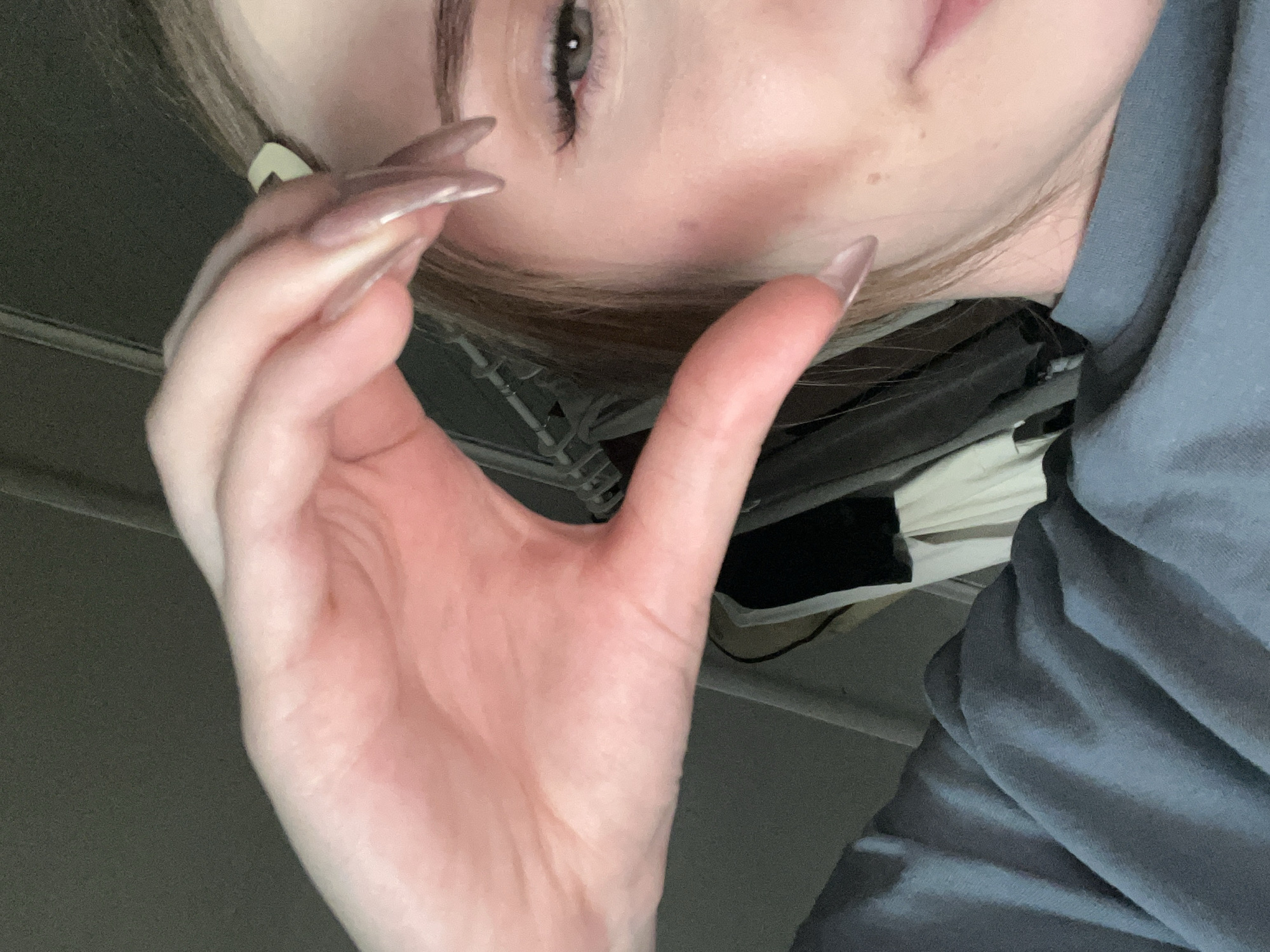 A close-up, indoor, waist-up, eye-level shot shows a young woman's right side, with her right hand held up to her face forming a heart shape. Her hand is positioned over her right cheek and eye, with her long, almond-shaped, light-pink-colored fingernails visible. She is wearing a blue, long-sleeved shirt. Her hair is blonde and pulled back, with a light-colored hair clip visible. The background is out of focus and appears to be an indoor setting with clothing racks in the distance. There is no indication of the city of Ashkadar, Russia, or any specific urban environmental features.