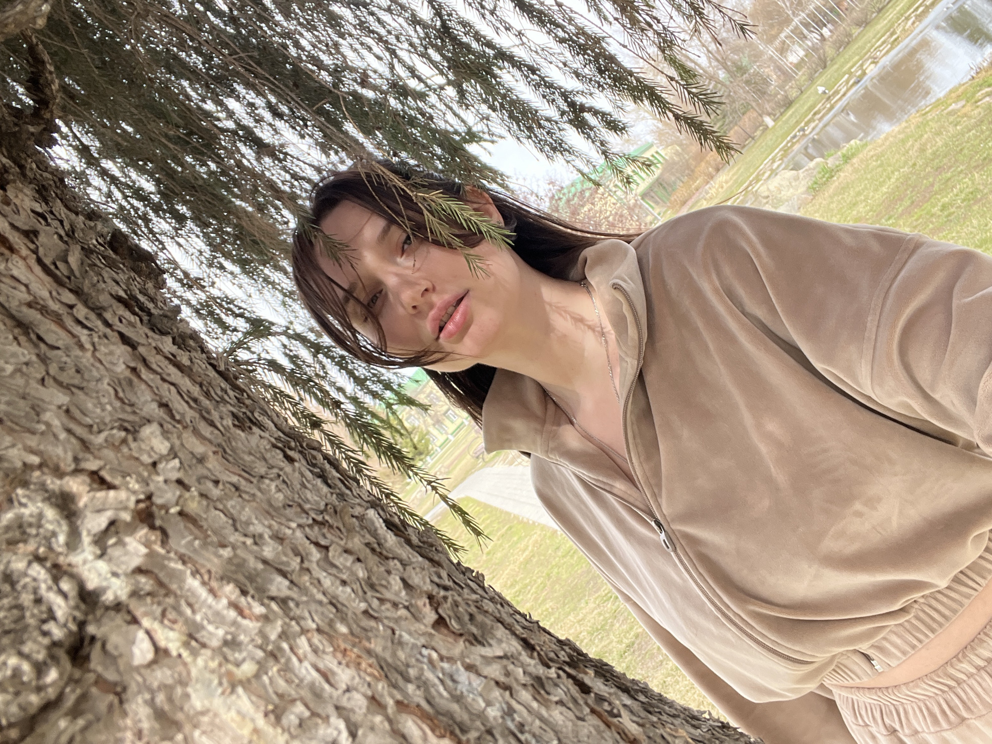 A young woman stands next to a tree in a park. She is wearing a beige zippered jacket and matching pants. Her dark hair is partially obscured by pine needles. In the background, there is a pond and a small building. The location is likely in the city of Kargopolye, Russia.
