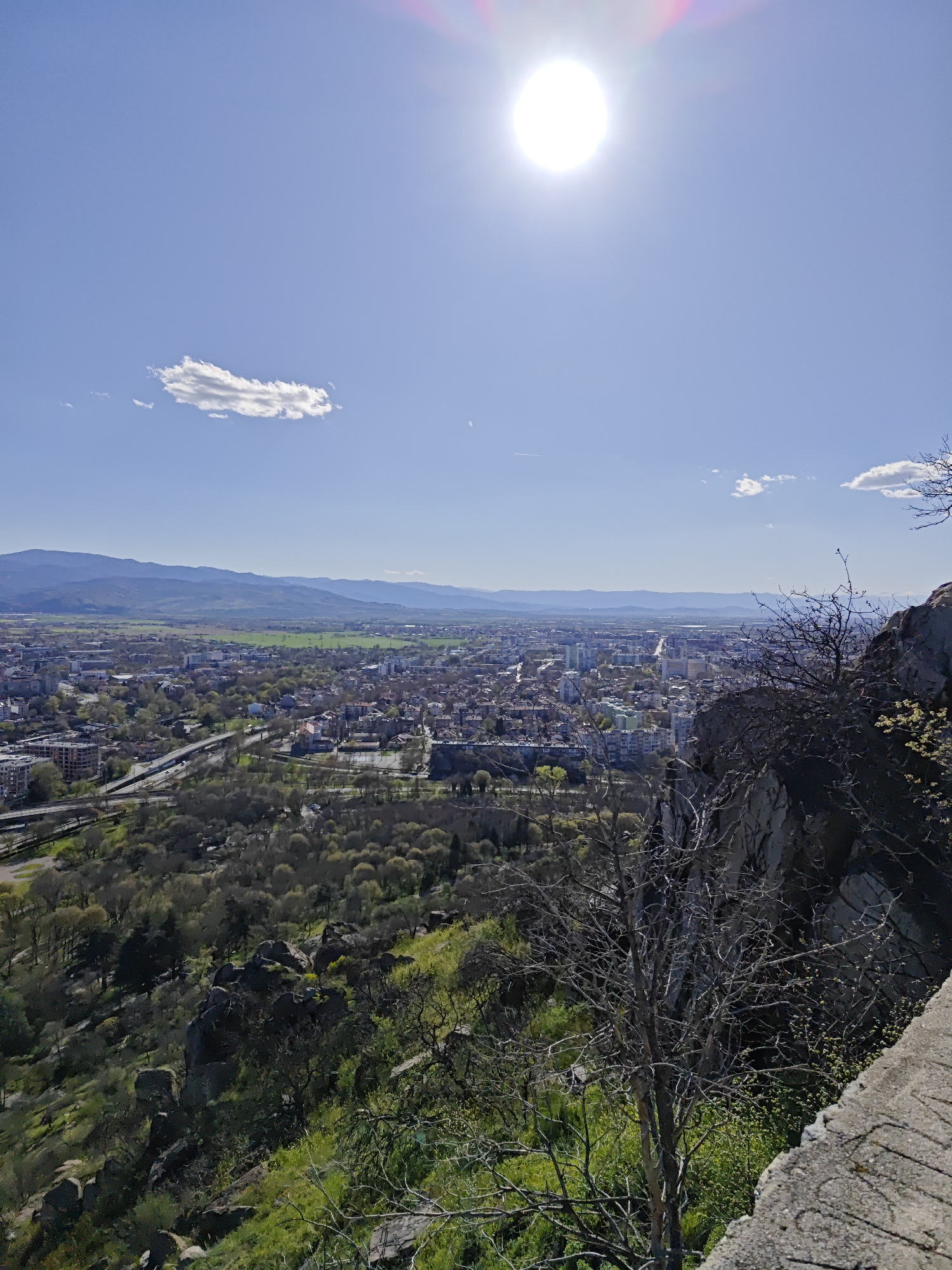 A high-angle, wide shot captures an expansive view of Plovdiv, Bulgaria, under a bright, clear sky with a radiant sun at the top of the frame. Scattered clouds dot the sky. The city stretches from the foreground to the distant mountains that form the horizon. The foreground consists of rocky terrain covered with green vegetation and bare trees. A stone ledge marks a boundary in the lower right corner. The midground transitions to the cityscape, revealing buildings, roads, and patches of greenery. A highway-like structure is seen winding through the city. The background features a range of mountains in the distance. The overall scene suggests a sunny, daytime view from an elevated vantage point.