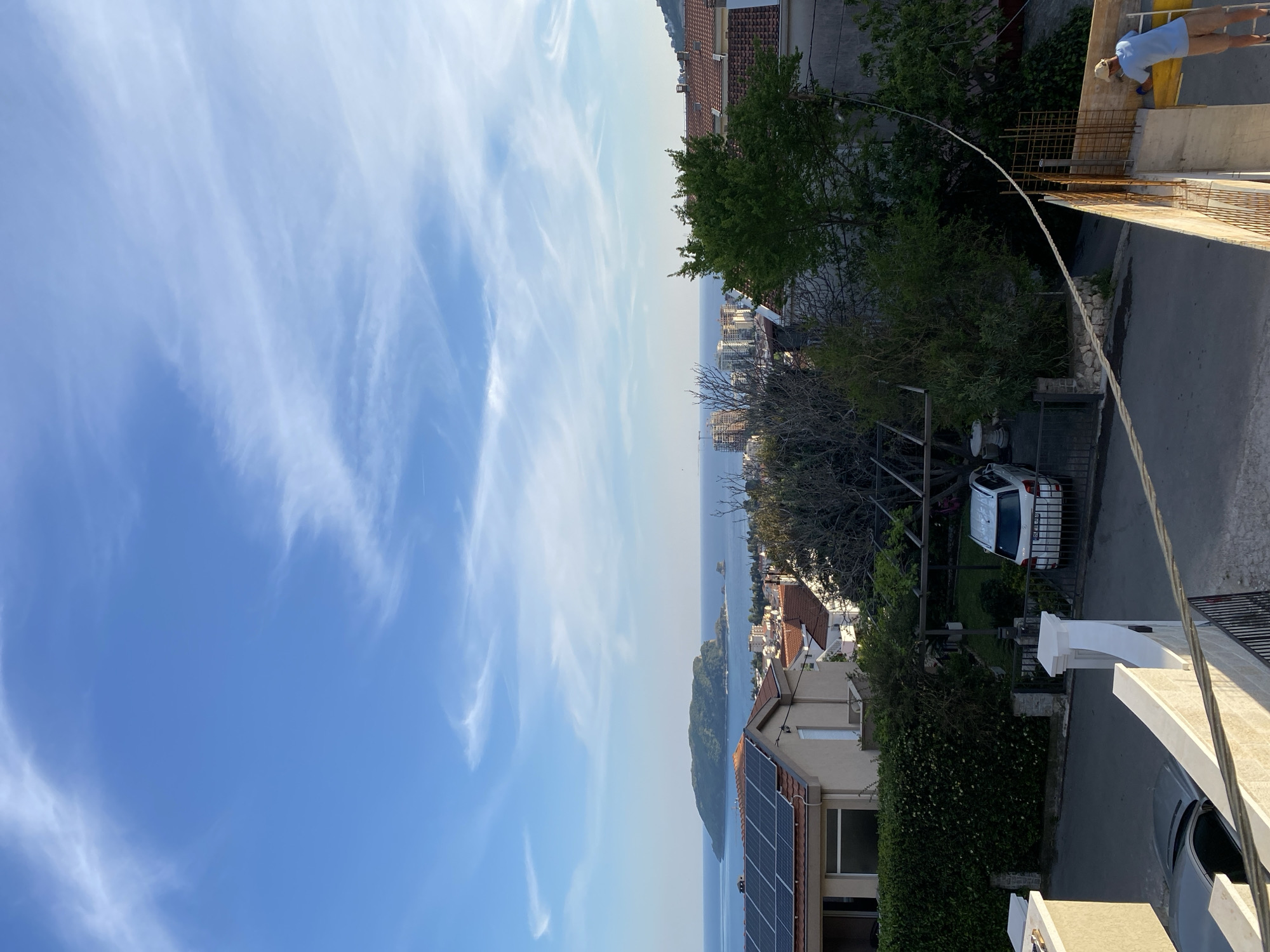 The image is a slightly elevated, oblique view of a coastal scene in Budva, Montenegro. The sky is predominantly blue with wispy cirrus clouds. In the middle ground, the Adriatic Sea stretches to the horizon, dotted with small islands and the coastline. The foreground is dominated by residential buildings with red-tiled roofs, solar panels and lush greenery, including trees and hedges. A white car is parked near one of the houses. An electrical cable stretches into the foreground where a construction worker is bent over working on a new construction. A portion of a gray vehicle is visible in the lower right corner. The scene is bathed in bright daylight.