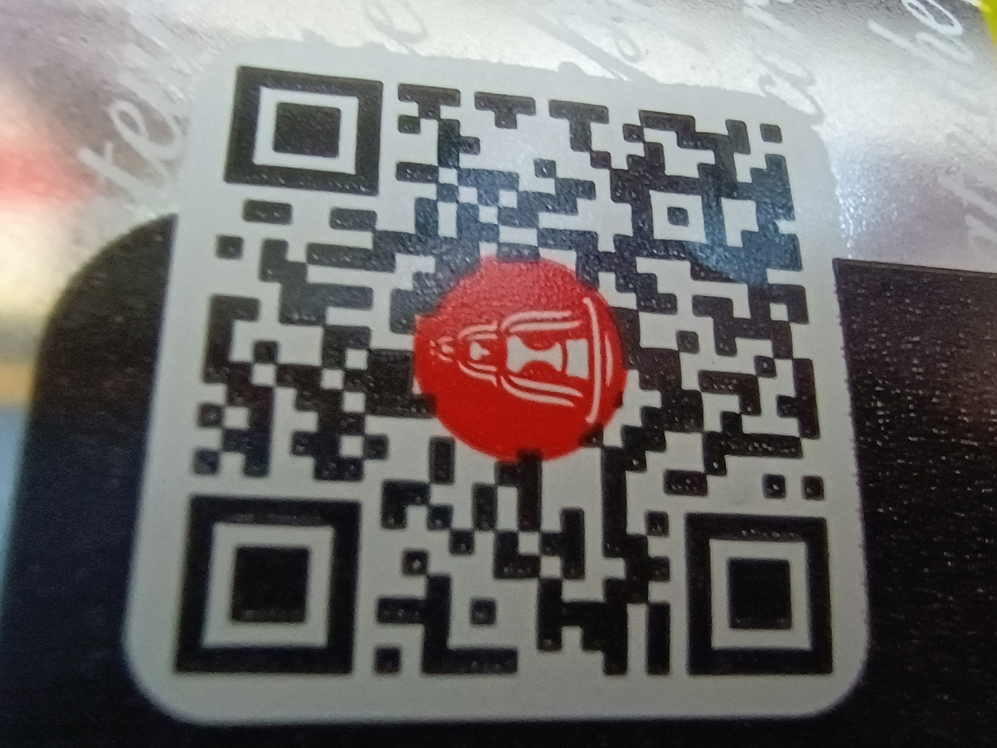 The image is a close-up shot of a QR code on a white sticker, which is partially affixed to a textured, possibly metallic, surface. The QR code is mostly black and white, with a red circular emblem overlaying a portion of its center. The emblem contains a stylized white design, possibly a logo or icon. In the background, faint white cursive text is visible on the textured surface, with some letters suggesting words like "tel". The overall lighting suggests an indoor or overcast environment. The image appears to be taken in Zaporizhzhia, Ukraine.