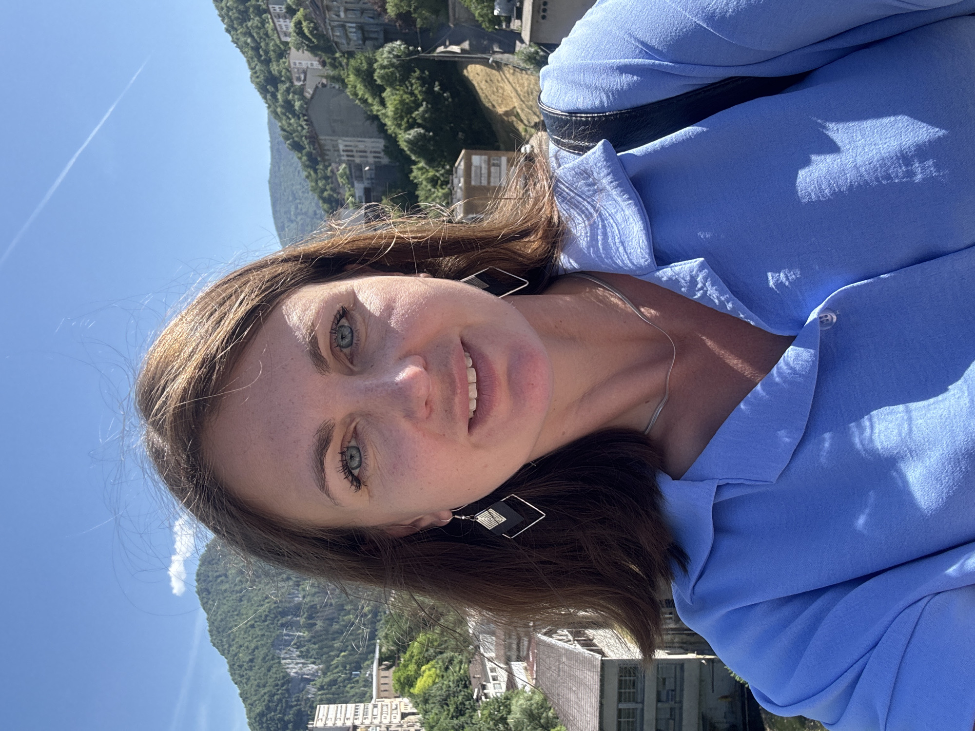 In this selfie taken outdoors, a woman with shoulder-length brown hair smiles at the camera. She is wearing a blue collared shirt and diamond-shaped earrings. Her eyes are blue-green, and her skin is lightly tanned. The background shows a town nestled in a valley with buildings and lush green mountains under a clear blue sky. This appears to be the city of Saint-Claude, France.
