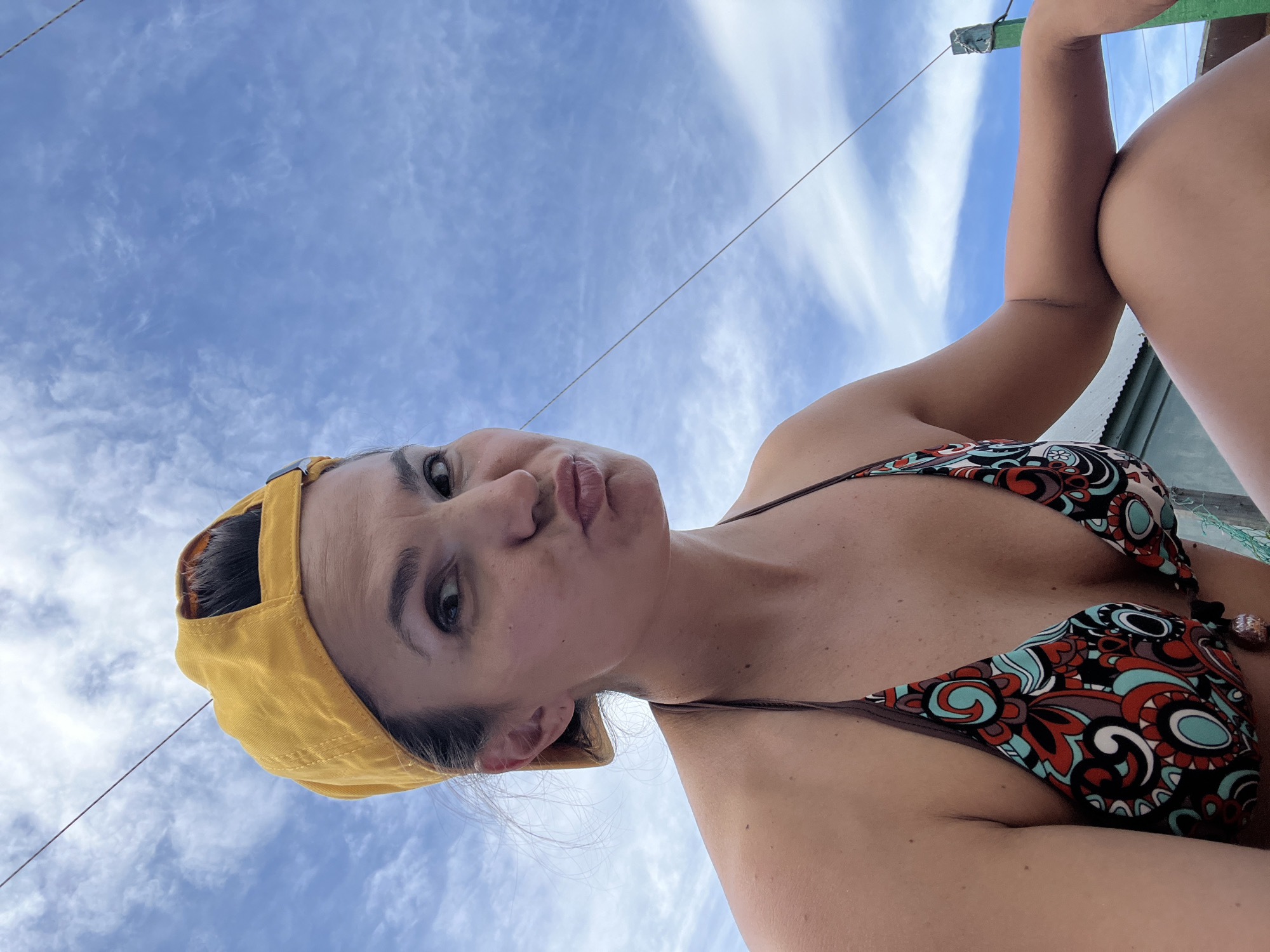 A woman wearing a yellow baseball cap and a patterned bikini poses for a selfie outdoors, with a bright blue sky filled with wispy clouds in the background. She is looking directly at the camera with a playful pout. The image is taken from a low angle, with the sky occupying the majority of the frame. The scene does not contain any discernible elements that would place it specifically in Petropavlovsk-Kamchatsky, Russia.