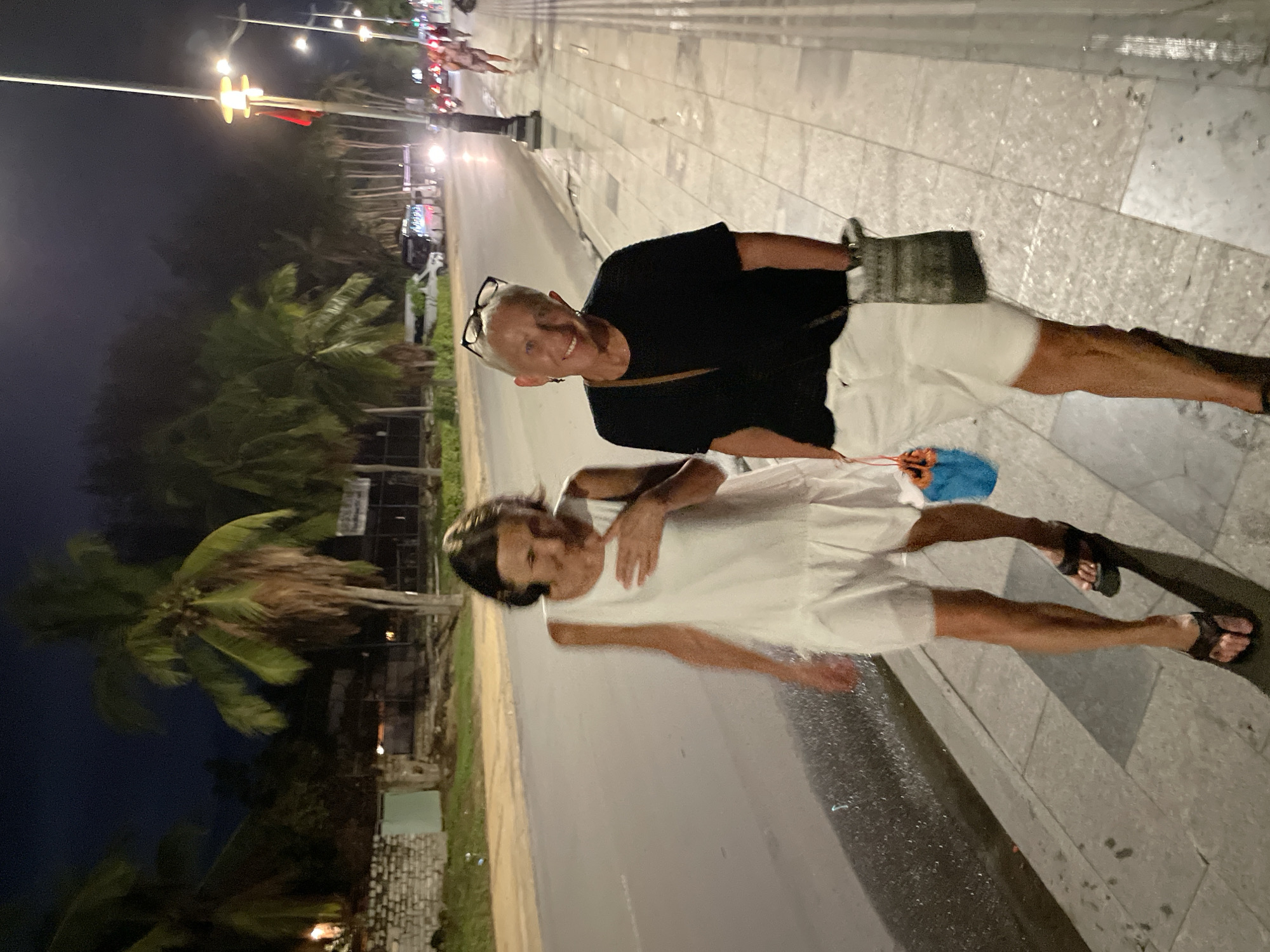Two people stand on a sidewalk next to a road in Phu Thuy Ward, Vietnam. One person wears a black shirt and white shorts with a small bag. The other person wears a white dress. The sidewalk is made of light-colored tiles, and the road is dark. Palm trees and street lights are visible in the background. Cars and motorcycles are on the road. The scene appears to be at night.