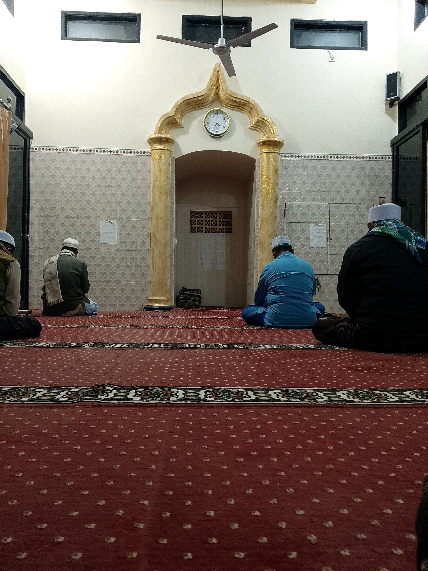 Four men are seated on a red carpet with a repeating pattern of small, light-colored floral motifs. The men are facing away from the camera, towards a mosque entrance that features ornate golden pillars and an arched entryway. Above the entrance, a round clock with Roman numerals is visible on the wall. The room appears to be indoors, with white walls and tiled sections. A ceiling fan is visible in the upper portion of the image. The overall setting suggests a religious service or prayer in a mosque. The location is identified as Rejoyoso, Indonesia.