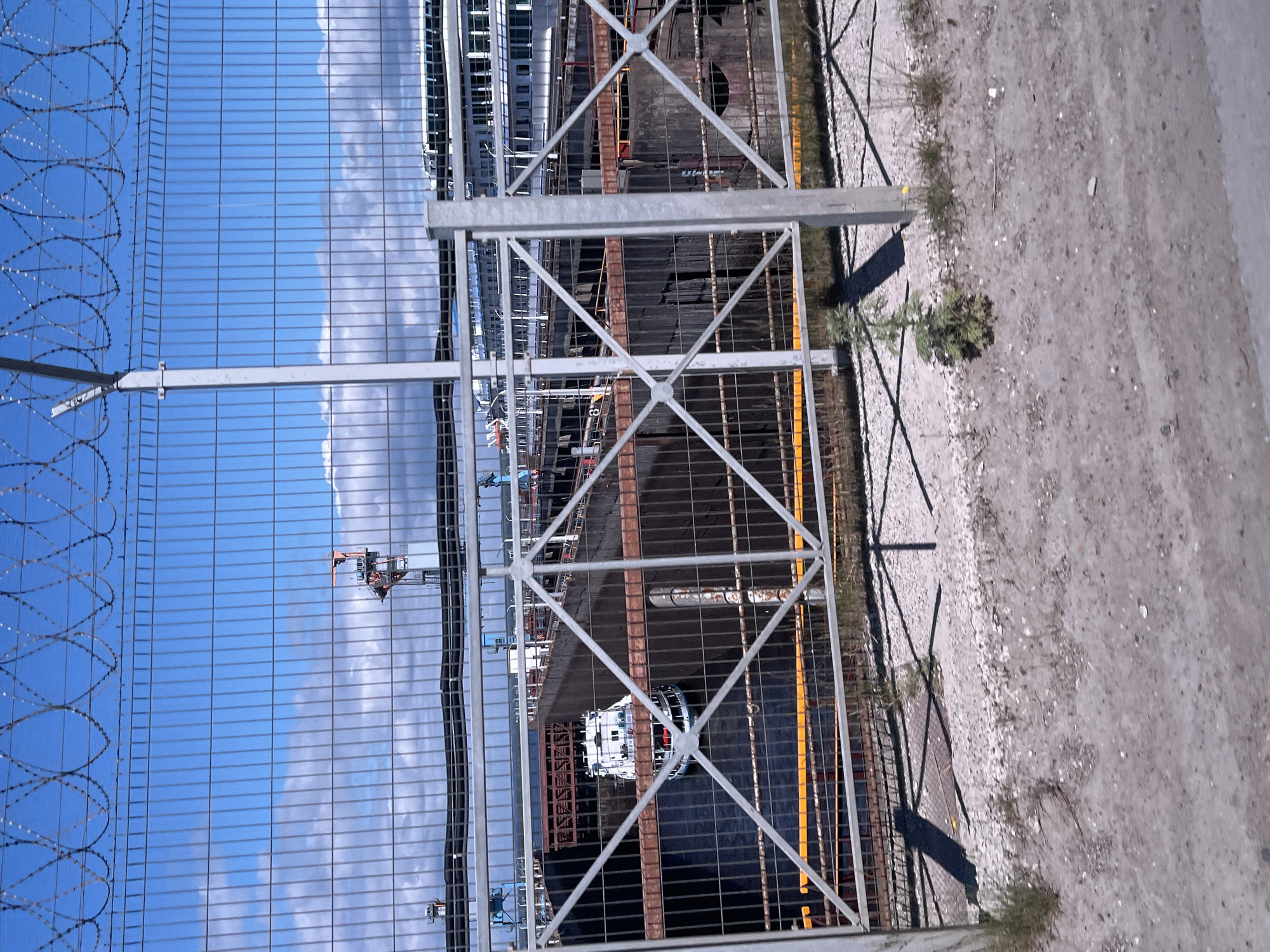 This image shows a view through a chain-link fence with barbed wire on top. The fence encloses an industrial area, likely a port or shipyard, in the city of Chaikovsky, Russia. In the foreground, the ground is covered with gravel and dirt, with some sparse vegetation. Behind the fence, a large, dark-hulled vessel is docked in a body of water. The vessel appears to be a barge or a freighter. Further in the background, there are cranes, industrial structures, and a large white cruise ship docked. The sky is blue with some clouds. The scene appears to be a typical day at a port.