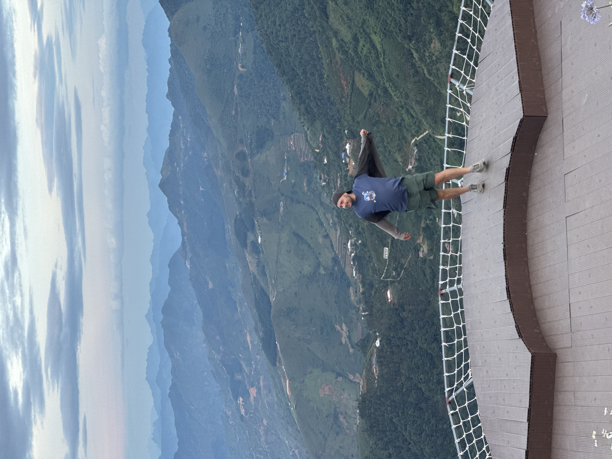 A Caucasian male stands centrally on a dark brown, multi-level composite deck platform at a scenic overlook in Ta Xua, Vietnam. He is dressed in a dark beanie, a dark blue t-shirt with a light blue graphic, dark green shorts, and grey athletic shoes. A dark grey long-sleeved garment is worn over his t-shirt, with sleeves creating a wing-like effect as his arms are outstretched to the sides, slightly bent at the elbows. He gazes towards the left with a slight smile. A white safety net with a grid pattern lines the curved edge of the platform.
Beyond the platform, a vast expanse of verdant mountainous terrain stretches into the distance under a cloudy sky. The closer mountains are densely covered in green foliage, revealing winding dirt roads and scattered small buildings or settlements on their slopes and in the valleys. As the mountains recede into the background, they take on progressively lighter and bluer hues due to atmospheric perspective, culminating in a faint blue ridge line under a sky filled with layered grey and light blue clouds. The overall lighting is soft and diffused. Small white and purple flowers are visible on the lower edges of the platform.