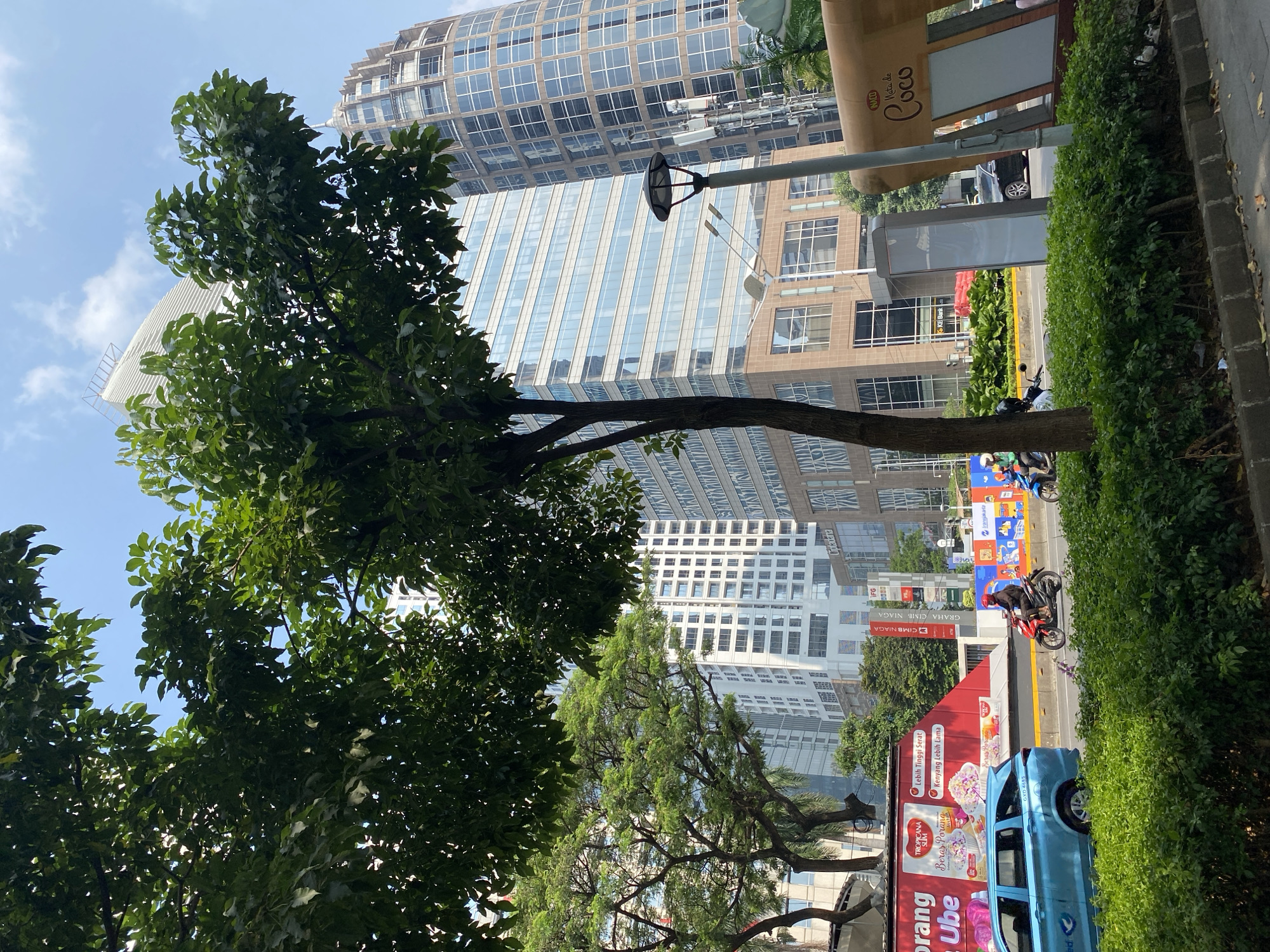 In South Jakarta, Indonesia, a street scene unfolds with urban greenery and modern architecture under a clear blue sky. The foreground is dominated by lush, manicured hedges lining the sidewalk, with a tree casting shadows on the road.
Vehicles are present, including a light blue minivan and several motorcycles. On one motorcycle, a person wearing a red helmet is visible, indicating movement along the street. Another motorcycle further down the road also has a rider.
Towering buildings form the backdrop, showcasing contemporary city design. One building is a tall, rectangular structure with numerous windows, while another is a curved, cylindrical building with a distinctive facade. Billboards and signage are present along the street, one featuring an advertisement for "Tropicana Slim" and another with text in Indonesian. Streetlights are also visible, one with a unique, modern design.
The overall atmosphere is one of a bustling urban environment with a touch of natural elements.