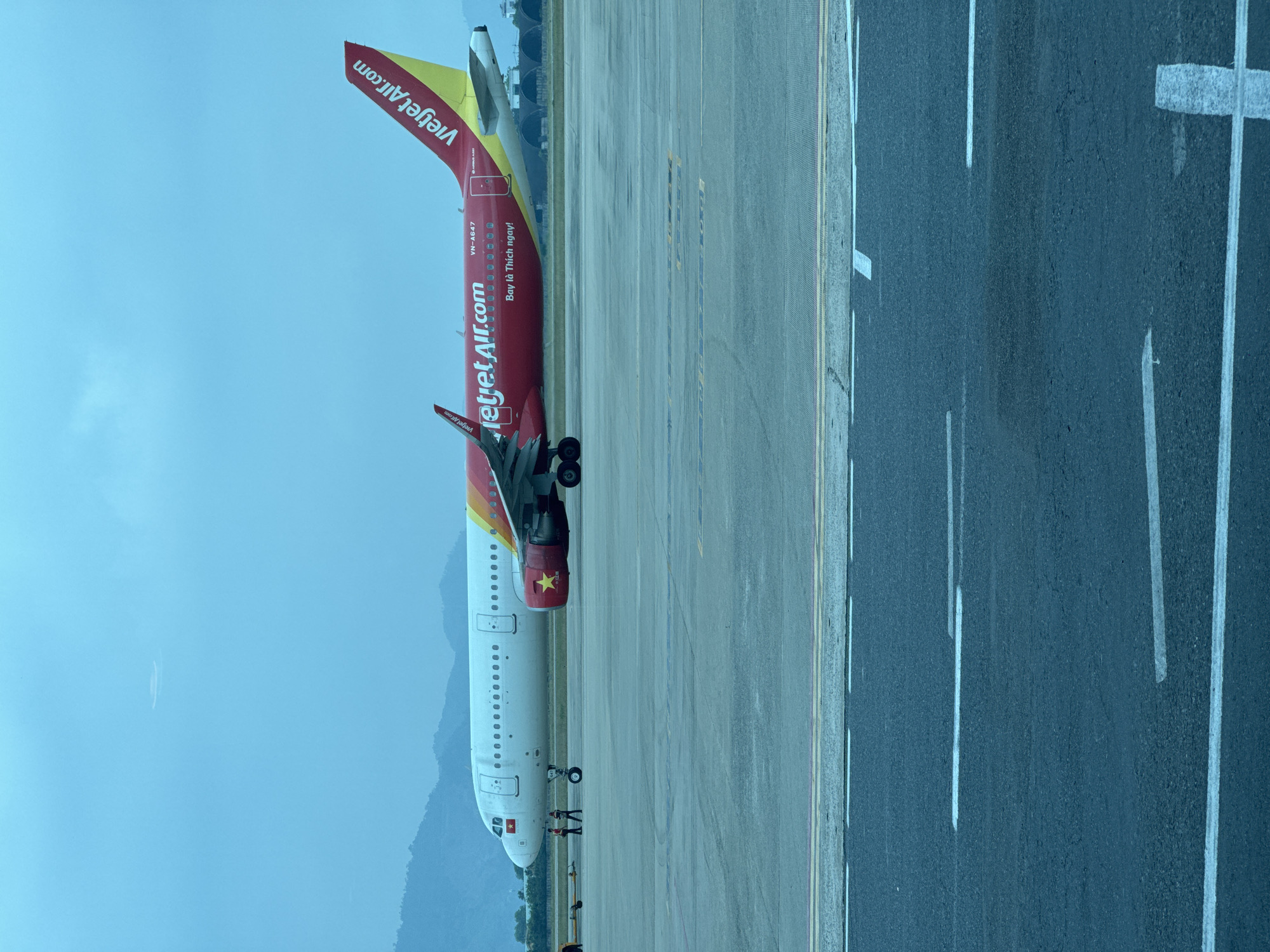 A red and yellow Vietjet Air airplane is parked on an airport tarmac. The plane's tail features the "VietjetAir.com" logo. The tarmac has visible markings and lines. The background includes a hazy sky, mountains, and hints of vegetation. The location is presumably Đà Nẵng International Airport in Vietnam. There are a few individuals visible near the front of the plane.