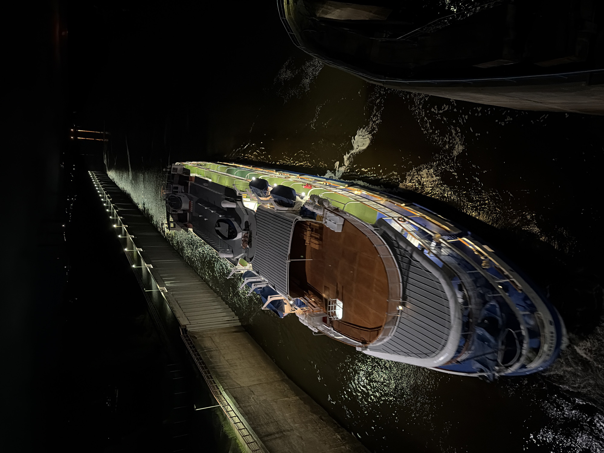 A multi-deck passenger vessel, identified by the inscription 'БЕРГЕН' (BERGEN) on its bow, is depicted navigating a narrow, concrete-walled waterway at night. The vessel, featuring a white and blue hull with green and brown upper deck sections, is brightly illuminated by internal lights spilling from its windows and by external deck lighting. A white wake is visible at the bow and along the port side, indicating forward motion.
On the viewer's left, a long, straight concrete retaining wall forms the boundary of the channel. This wall is topped with a series of uniformly spaced pole-mounted lamps that project light onto the water surface and the wall itself. A metal railing runs along the upper edge of this wall, and a smaller, unlit concrete barrier is visible further left, parallel to the main wall.
On the viewer's right, another concrete structure forms the opposing boundary of the waterway. This wall appears darker due to less direct illumination. The water within the channel is dark, reflecting the artificial lights from the vessel and the shore-side lamps, creating patterns of illuminated ripples.
The overall environment is characterized by profound darkness, with only faint, distant artificial lights visible beyond the immediate channel. This scene is situated in the city of Chaikovsky, Russia, and is consistent with a navigational lock or canal system, specifically the Votkinsk Dam's lock on the Kama River. No individuals are discernible on the vessel or along the channel walls.