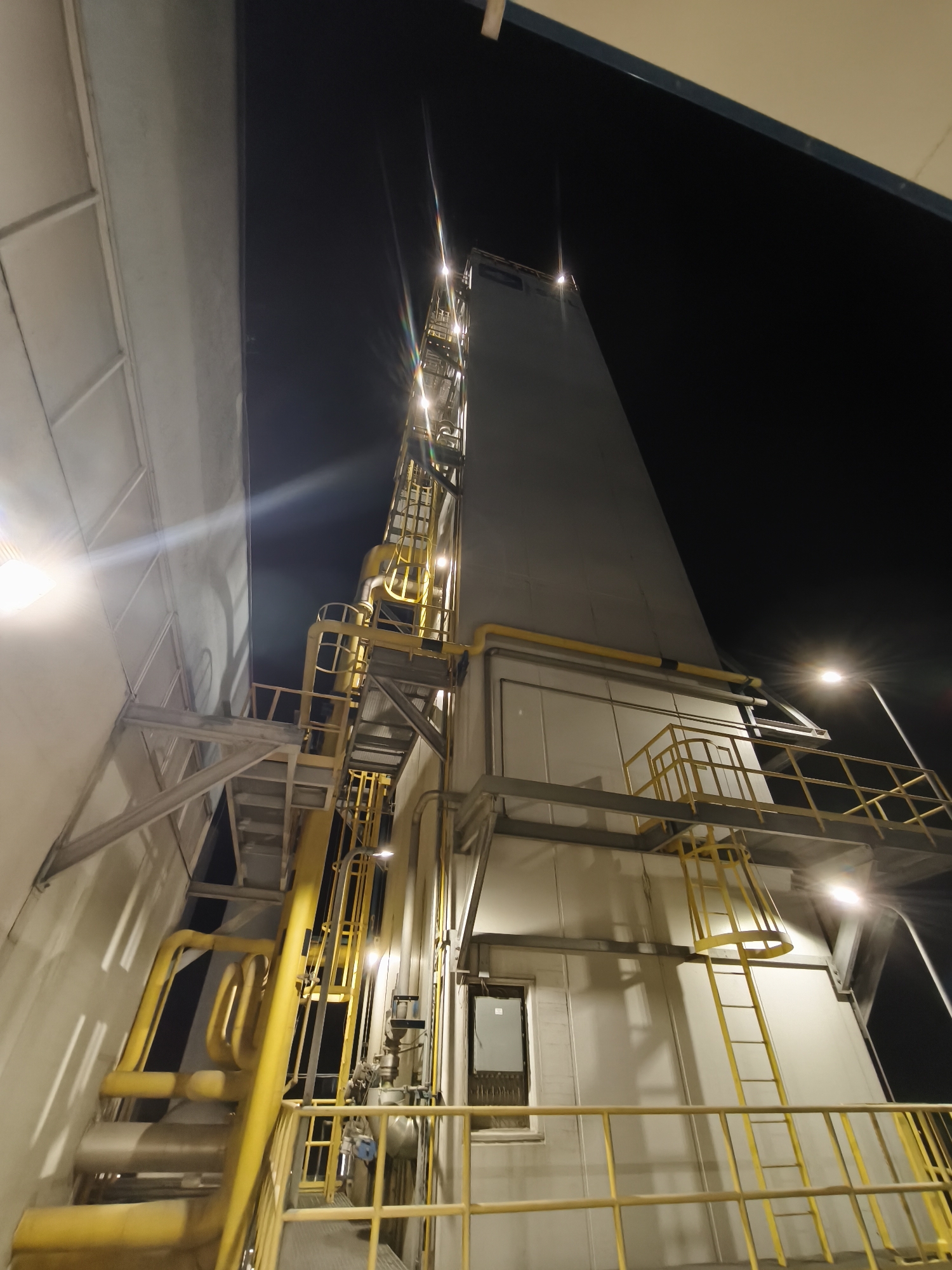 The image captures an industrial scene at night. A tall, gray structure dominates the view, featuring yellow piping and ladder systems ascending its height. Illumination is provided by several lights, both on the structure and from surrounding sources, creating highlights and shadows. The structure is adjacent to a building with a light-colored facade. The sky is dark, suggesting the absence of daylight. The overall impression is of an industrial plant operating under artificial light. There are no people visible in the image. The location is in the city of Unknown, Unknown.