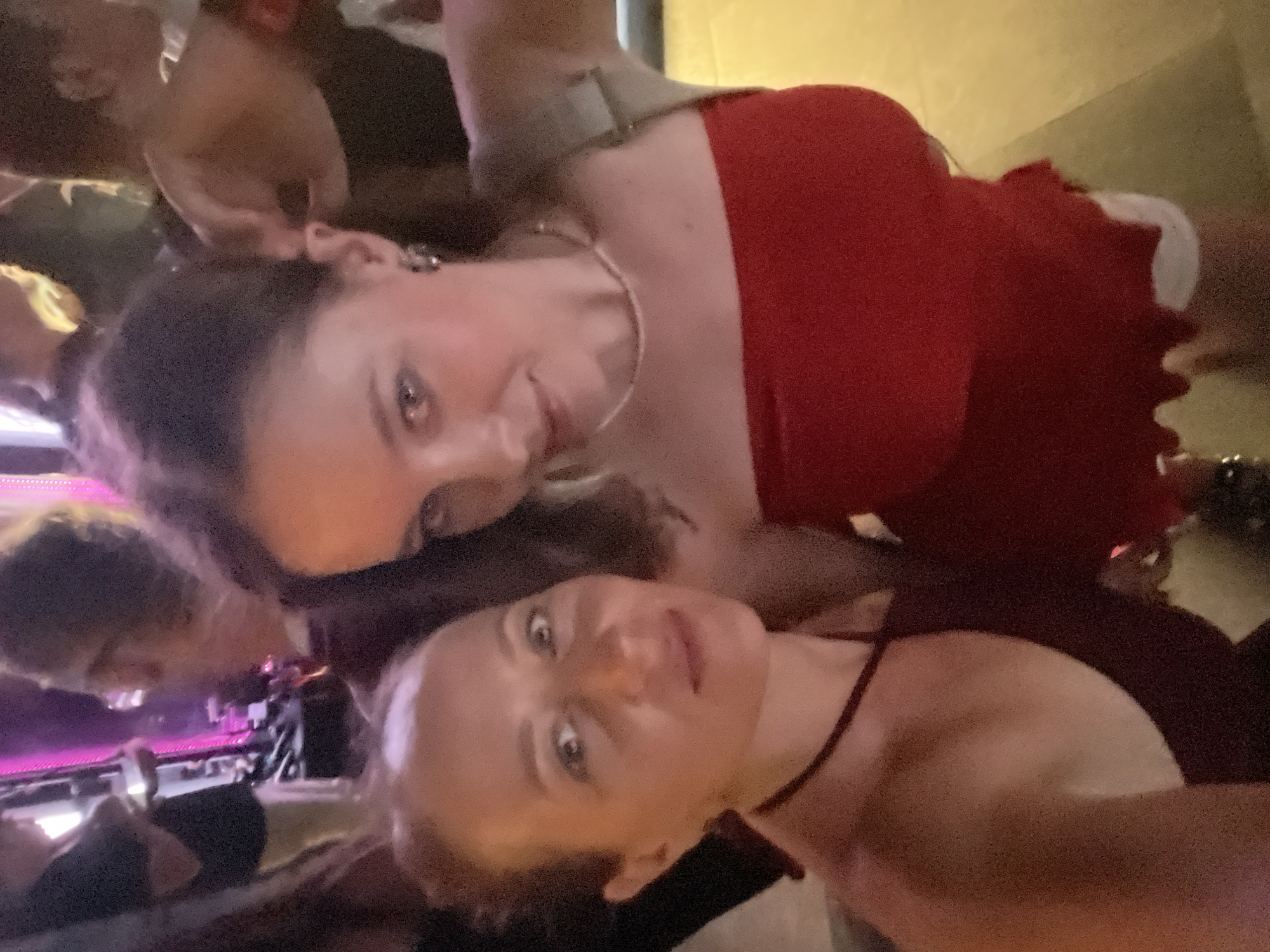 The media file captures a group of people in what appears to be a dimly lit indoor setting, possibly a bar or club, given the presence of colorful lighting in the background that suggests a stage or dance floor. Two women are prominently featured in the foreground, leaning towards the camera as if taking a selfie.
The woman on the left is wearing a red dress and has her hair pulled back. She is smiling, and her eyes are open, looking directly at the camera. The woman on the right is also looking at the camera, with her eyes wide open and a slight smile. She is wearing a dark-colored top. The image is slightly blurry, suggesting movement or a low-light environment.
Other individuals are visible in the background, mostly out of focus, indicating a social gathering. The overall atmosphere seems lively and informal. The lighting is a mix of ambient indoor light and the purplish hues of the background illumination.
No visible text is present in the image. The location context provided is Da Nang, Vietnam, but there are no specific visual cues within the image that confirm this location, such as distinctive architecture, signage, or local attire. The time of day is likely evening or night due to the indoor lighting conditions. The emotions conveyed by the visible subjects are cheerful and engaged, consistent with a social event.