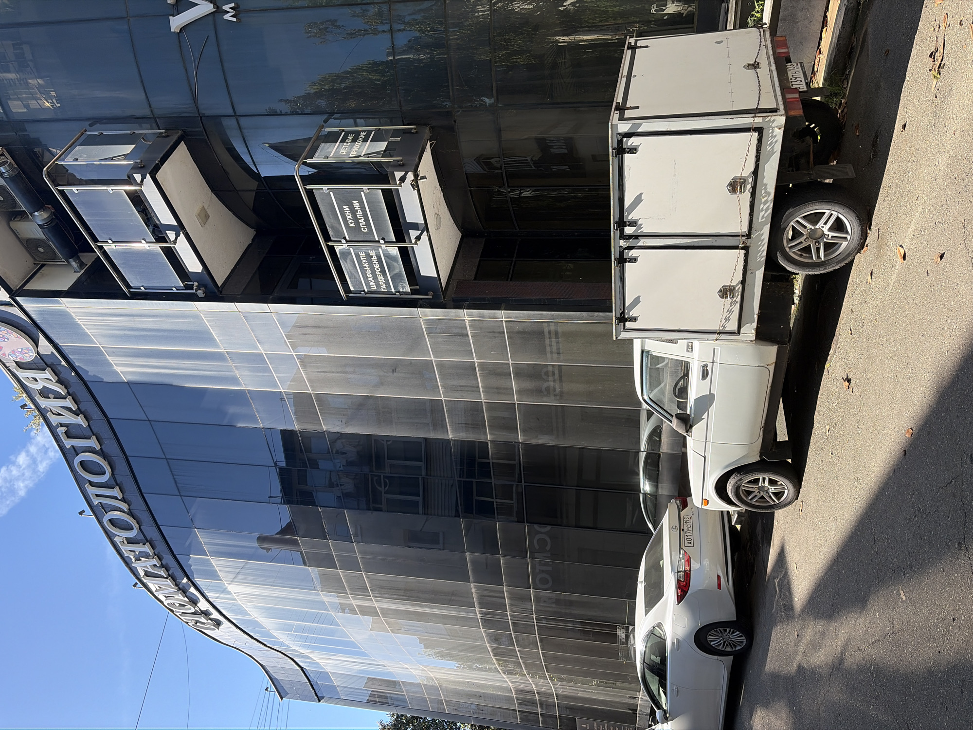 A street scene in Sochi, Russia, on a bright, sunny day, featuring a modern building with a dark, reflective glass facade and several parked white vehicles. The sky is clear blue, and sharp shadows are cast on the asphalt street, which has a few scattered dry leaves.
The building's curved upper edge displays a large sign with silver letters spelling "СТОМАТОЛОГИЯ" (Dentistry). Below, near what appears to be an entrance, smaller, multi-panel signs are mounted. These signs advertise various furniture or home interior services, including "ДЕТСКИЕ ПРИХОЖИЕ" (Children's Hallways/Entrances), "КУХНИ СПАЛЬНИ" (Kitchens, Bedrooms), and "ШКАФЫ-КУПЕ ГАРДЕРОБНЫЕ" (Sliding Wardrobes, Dressing Rooms). Reflections of trees and the sky are visible in the building's glass panels.
Two white cars are prominently parked along the curb. Closest to the viewer is a white Lexus sedan, bearing a Russian license plate "A017PC 193". Partially visible behind the sedan is another white car, a Lada Niva, identified by a "4x4" badge. Further down, a white utility truck with an enclosed box-style body and its rear doors open is parked. This truck displays a Russian license plate "739TH 123". No people or specific activity are visible in the image, suggesting a quiet moment on the street.