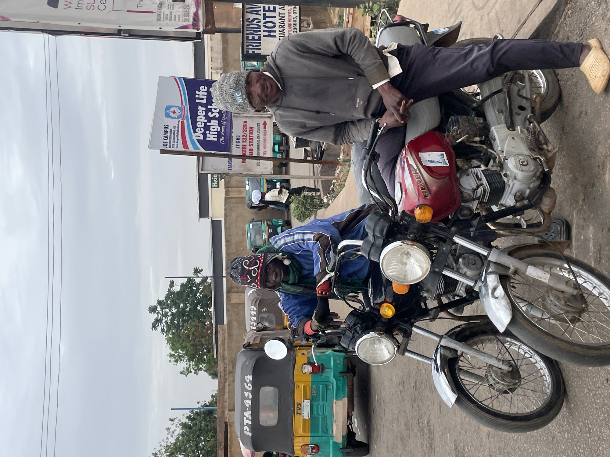 Two men sit on motorcycles parked on the side of a street in Jos, Nigeria. The man on the front motorcycle is wearing a blue shirt and a patterned hat. The man on the rear motorcycle wears a gray sweatshirt and a knitted cap. A yellow and green tricycle with the license plate "PTA-4564" is parked in front of the motorcycles. Behind the men and vehicles are street signs for "Deeper Life High School," "Friends Avenue Hotel," and a power sub-station. Buildings and trees are visible in the background.