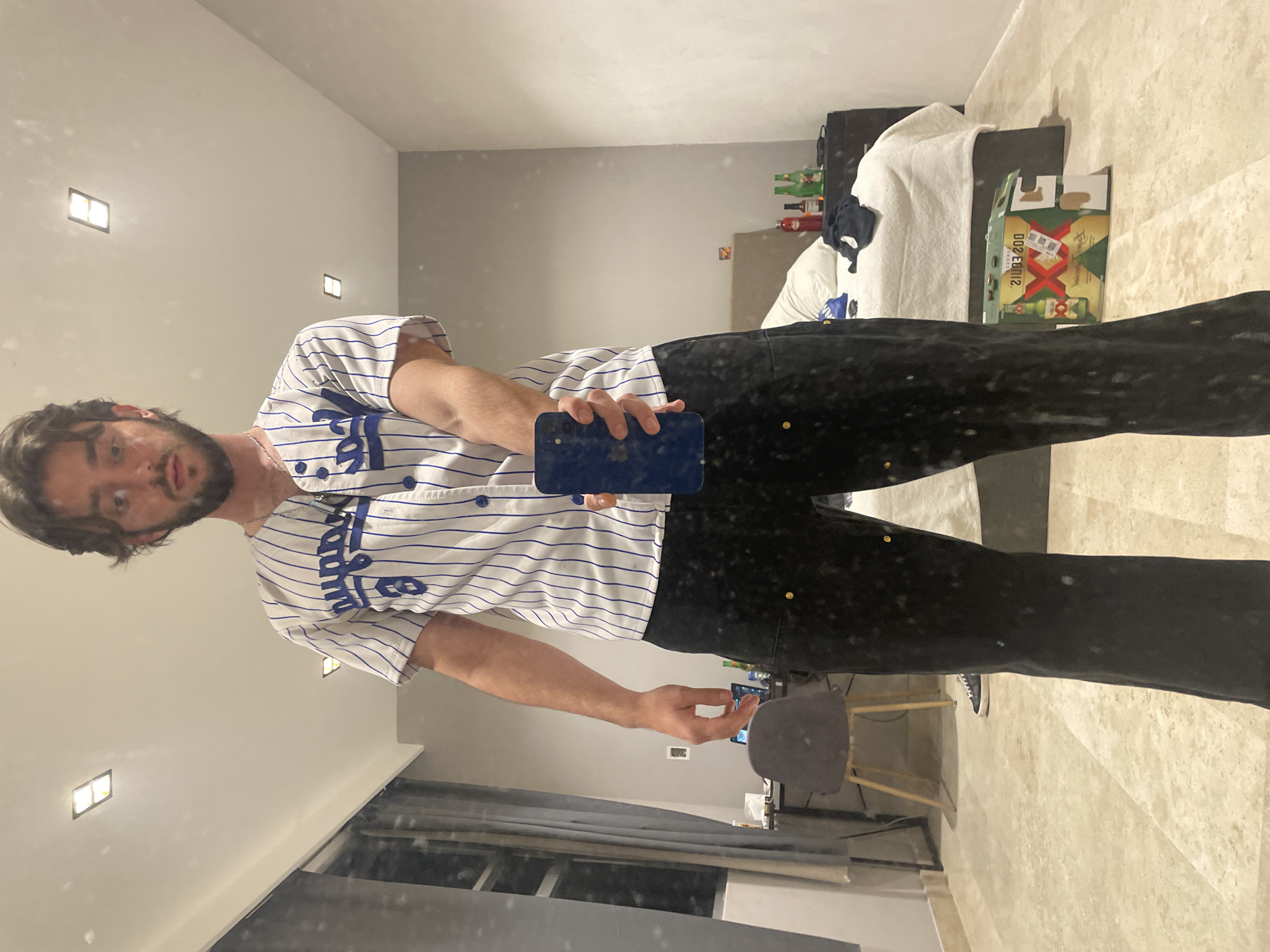 A young man stands in what appears to be a bedroom in a modern apartment in Huixquilucan de Degollado, Mexico. He is wearing a white pinstriped baseball jersey with blue lettering and buttons, and dark pants. He is holding a blue smartphone in his right hand, taking a selfie in a full-length mirror. The room is sparsely furnished, with a bed in the background, a desk and chair to the left, and a nightstand with bottles on it. A large cardboard box with what appears to be beer bottles is on the floor in the foreground. The lighting is dim, with recessed ceiling lights providing illumination. The overall impression is of a casual, personal moment captured in a domestic setting.