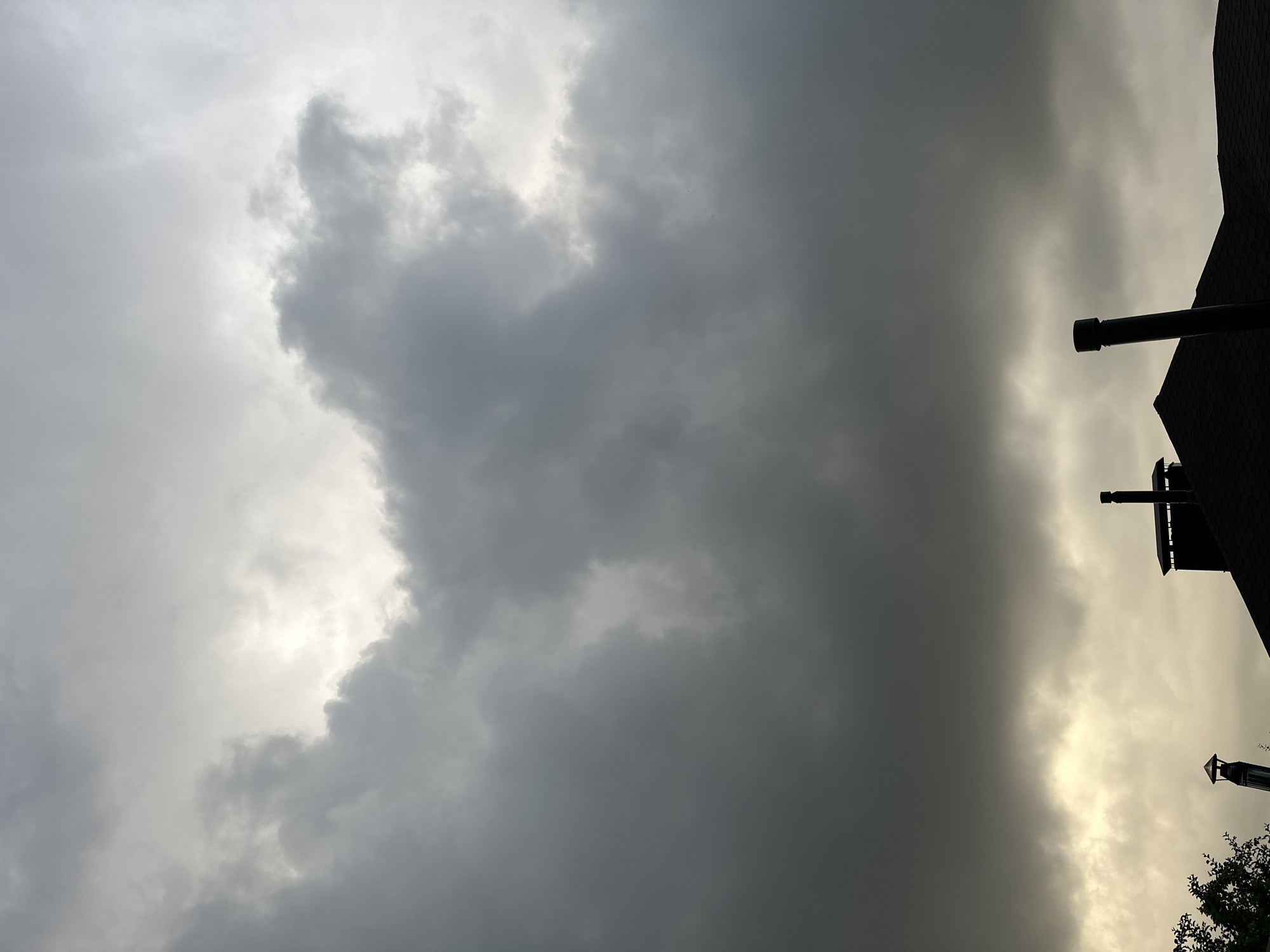 The image captures a dramatic sky over the city of Nizhny Novgorod, Russia. Dark, ominous storm clouds dominate the upper portion of the frame, with breaks in the clouds revealing a brighter, lighter sky beyond. The lower part of the image shows the silhouettes of rooftops with chimneys, suggesting an urban or suburban setting. The overall mood is somber and suggests an impending storm or the aftermath of one.