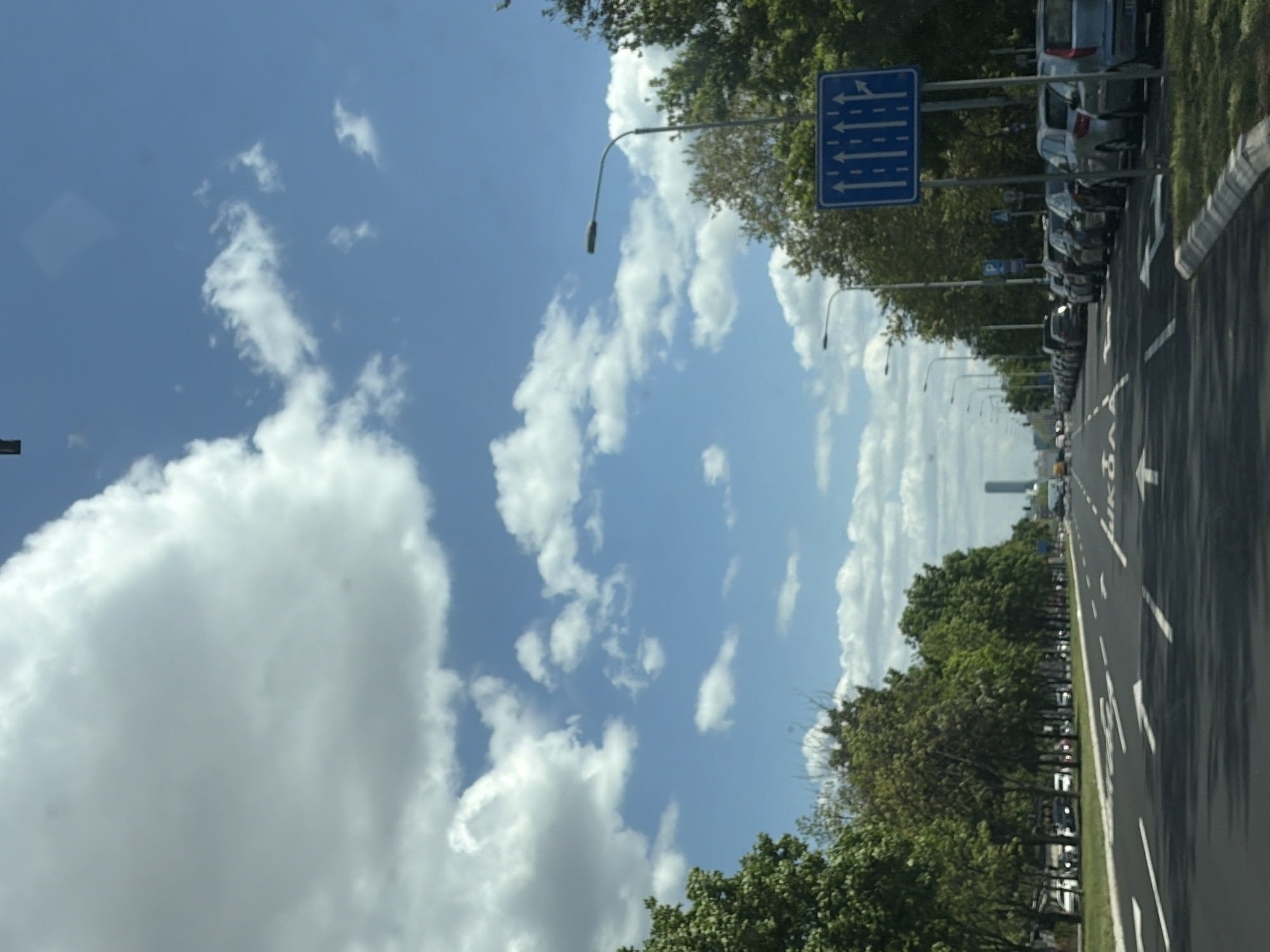 The image captures a street scene in Belgrade, Serbia. The sky occupies a significant portion, displaying a vibrant blue color dotted with cumulus clouds. A street runs parallel to the right side of the frame, featuring multiple lanes separated by white lines. Parked cars line the right edge of the road. Trees are planted along the roadside, adding a green border between the street and the sky. A blue road sign with white arrows indicates lane directions. Streetlights are positioned along the road. A tall building can be seen in the distance. There are no visible people in the image.