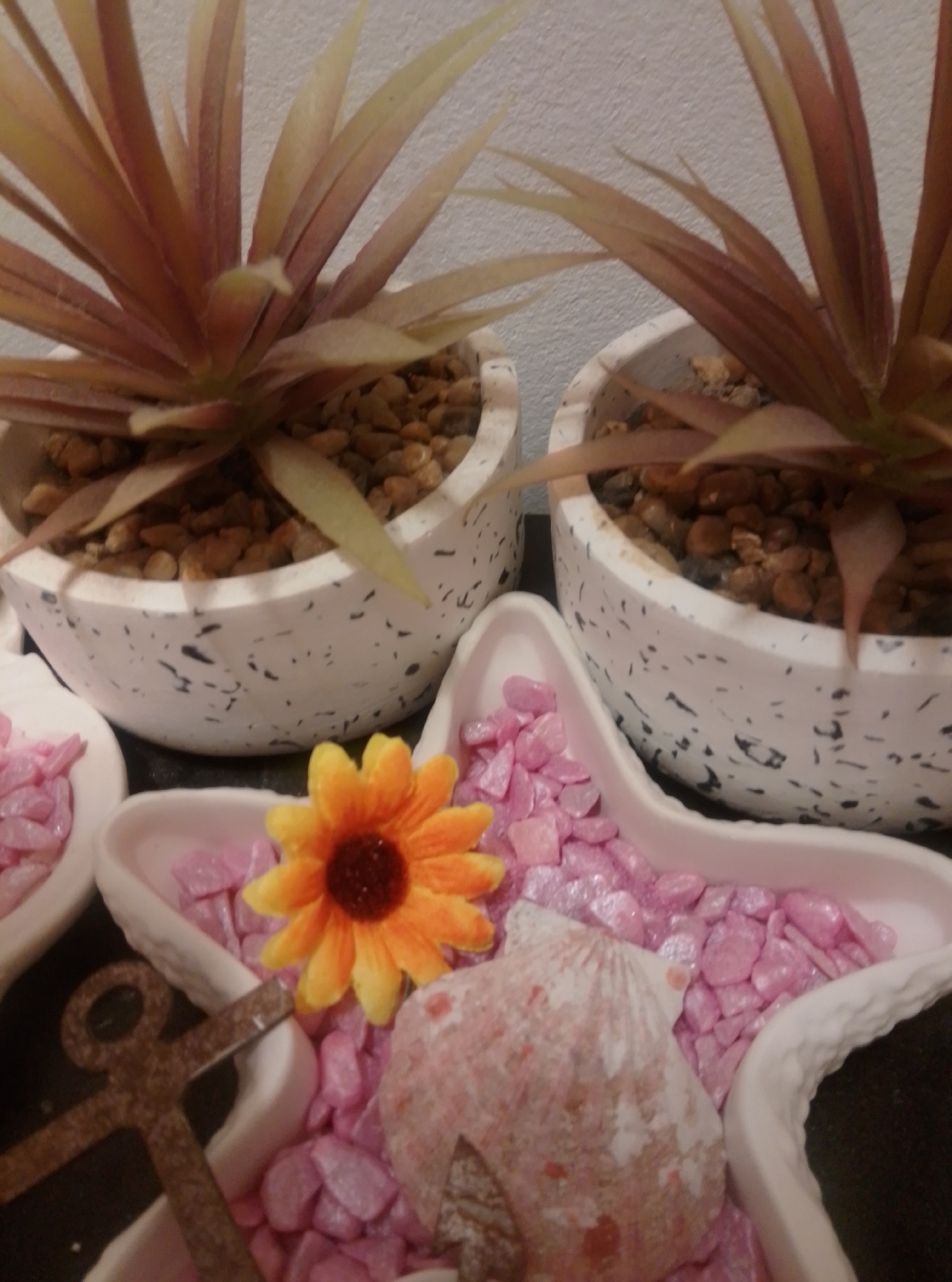 The image depicts a close-up, indoor scene featuring decorative items. Two potted plants with reddish-brown and pale green spiky foliage are visible in the background, planted in white speckled pots filled with small brown pebbles. In the foreground, a star-shaped white dish is filled with small pink gravel and decorative elements. A single, bright orange and yellow artificial sunflower is positioned in the center of the star dish. To the left of the sunflower, a rusty brown anchor-shaped ornament lies on its side. A large, pale pinkish-white seashell, also with some reddish-brown markings, is placed on top of the pink gravel in the lower right portion of the star dish. The background wall appears to be a light, textured grey. The lighting is dim, suggesting an indoor setting. No people are visible in the image. The image does not contain any elements that would indicate its location in Wedel, Germany.