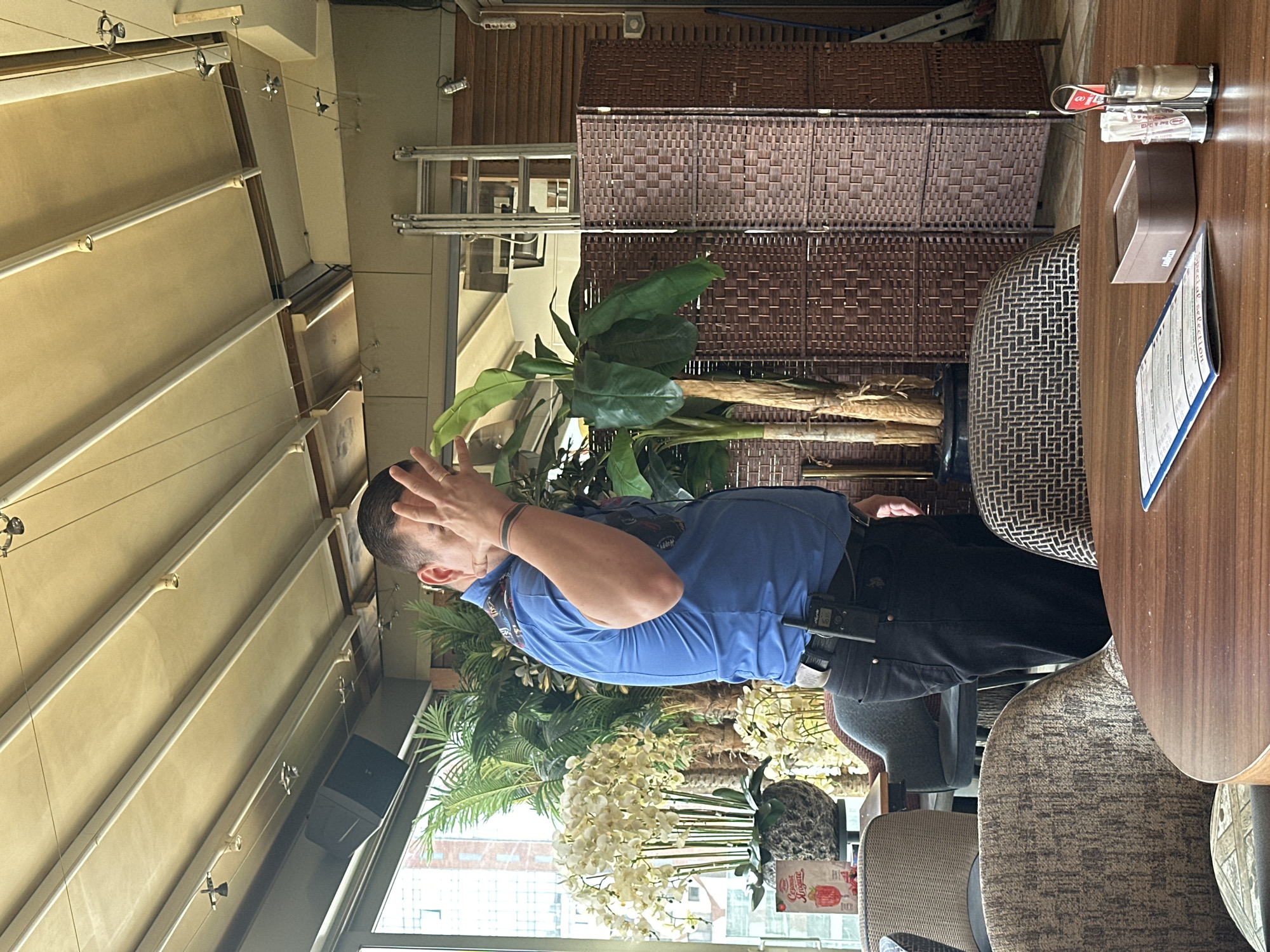 The image depicts a man in a blue polo shirt standing in what appears to be an indoor/outdoor cafe or restaurant space. The man is holding his hands near his head. He is wearing dark pants and has what appears to be a device or accessory attached to his belt. The setting features a partially covered ceiling, likely an awning or retractable roof. There are potted plants with white flowers and larger green plants in the background. A table with a menu, condiments, and a patterned chair are visible. Outside the window, there is a glimpse of a cityscape, which may indicate the location is in an urban area. Based on general knowledge and the location of buildings appearing through the window, the image is in the city of Sofia, Bulgaria.