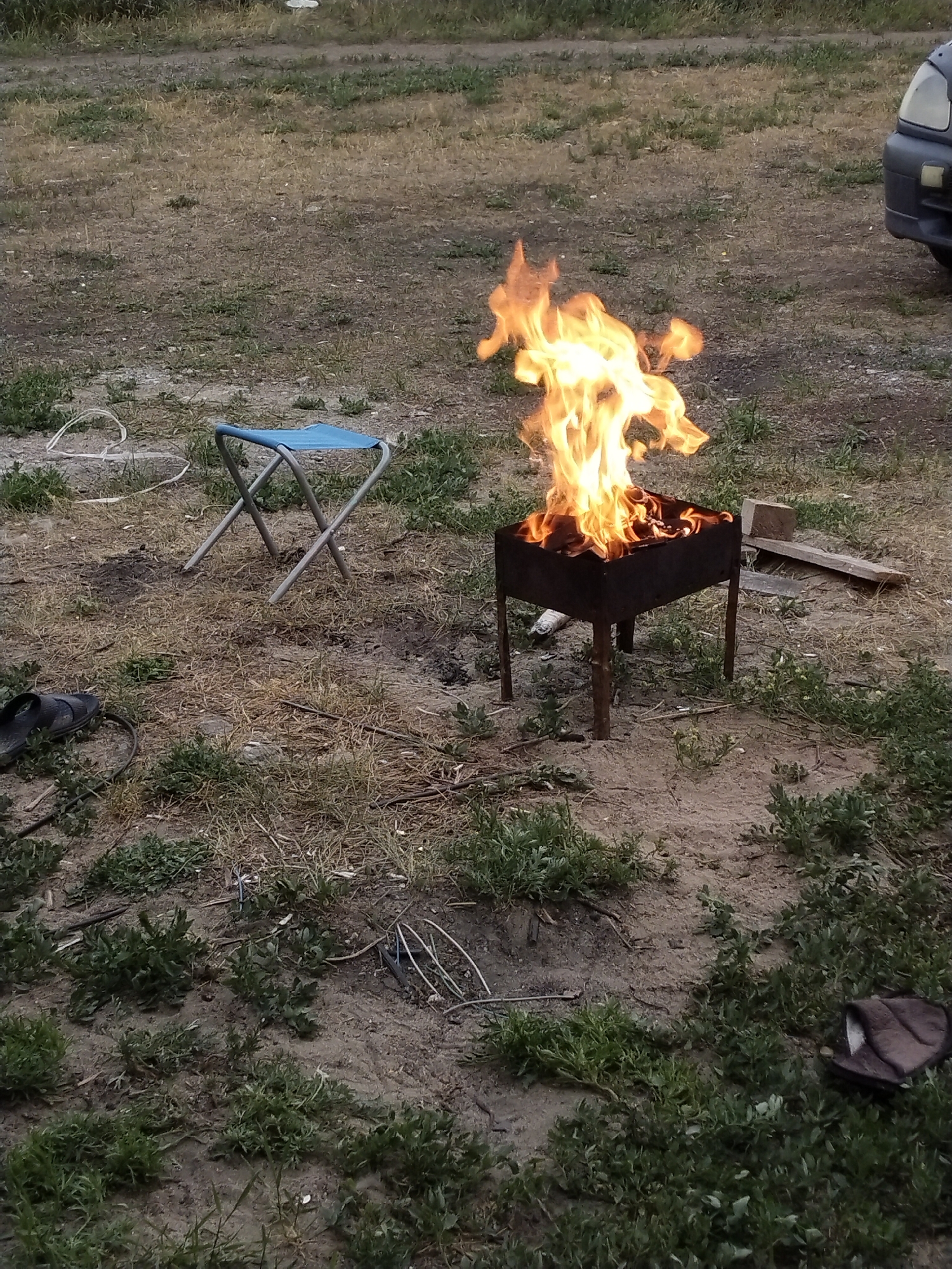 The image depicts an outdoor scene with a fire burning in a metal barbecue grill. Flames are prominently visible, reaching upwards from the grill. To the left of the grill, a folded blue camping stool is positioned on dry, uneven ground covered with sparse vegetation and dirt. In the foreground and scattered around the scene are patches of green grass and dry weeds.
To the left of the stool, a black sandal is visible. Further to the right, a light-colored wooden plank lies on the ground. In the background, a portion of a dark-colored vehicle is visible, specifically the front bumper and headlight area. The ground appears to be a mixture of sand, dirt, and dry grass.
The overall setting suggests an informal outdoor gathering or campsite. The lighting indicates it may be late afternoon or early evening, as the light is somewhat dim and the sky is not clearly visible. There are no people visible in the frame. The activity taking place is the preparation or presence of a fire for cooking or warmth. There is no visible text in the image. The location is identified as Зелёное, Russia.