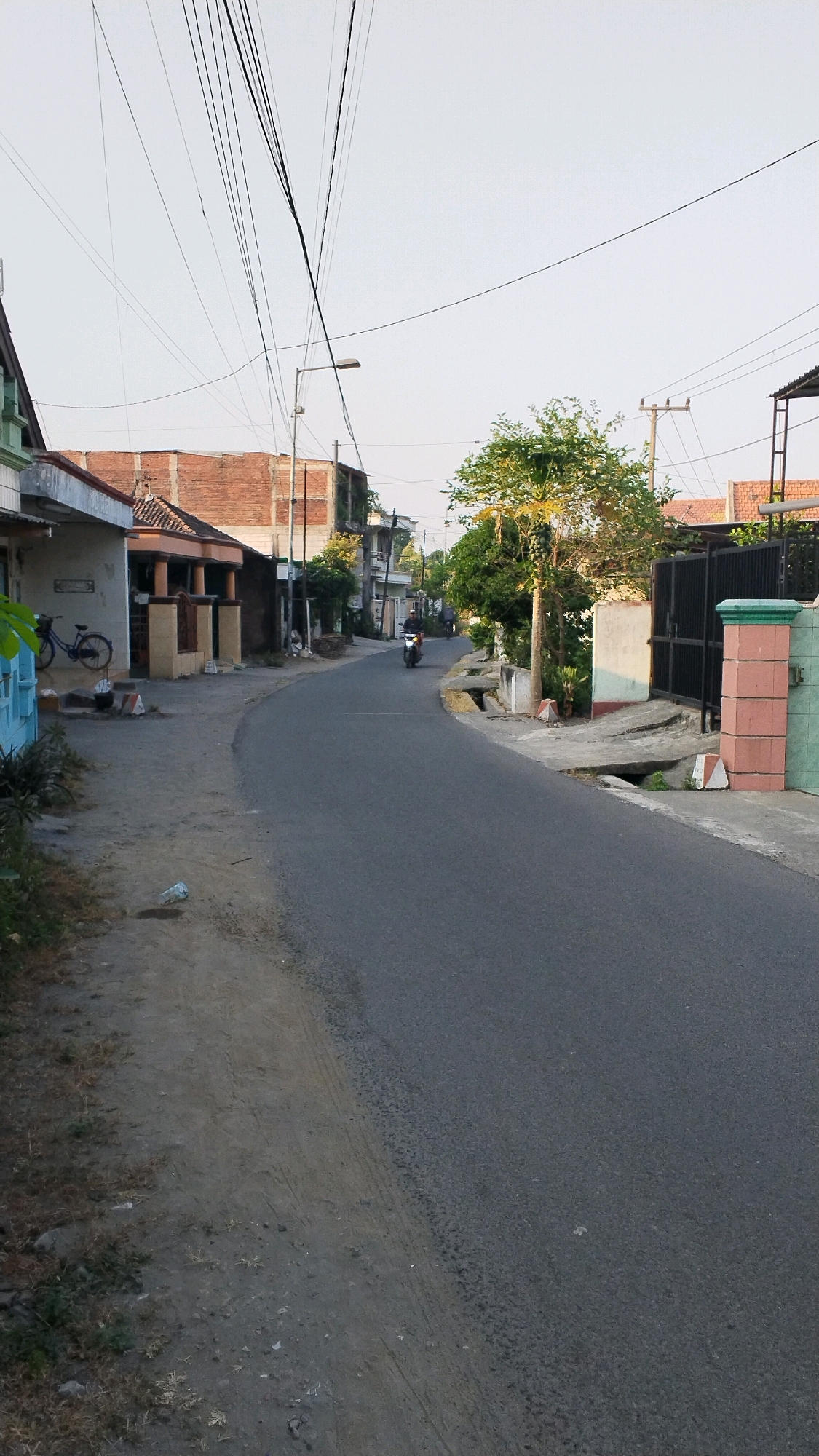 The image depicts a residential street in Penompo, Indonesia, likely during the late afternoon or early morning given the soft lighting. The asphalt road curves gently to the right. On the left side of the road, a row of modest houses with brick and painted walls are visible. A light blue bicycle is parked on the side of the road in front of one of the houses. Further down the street, two individuals are riding motorcycles.
The right side of the road features more houses, some with black metal gates and decorative pillars. There are several trees and vegetation along the sides of the street, adding a touch of green to the urban landscape. Overhead, a complex network of power lines crisscrosses the sky, a common sight in many Indonesian urban and semi-urban areas. The overall impression is one of a quiet, everyday street scene.