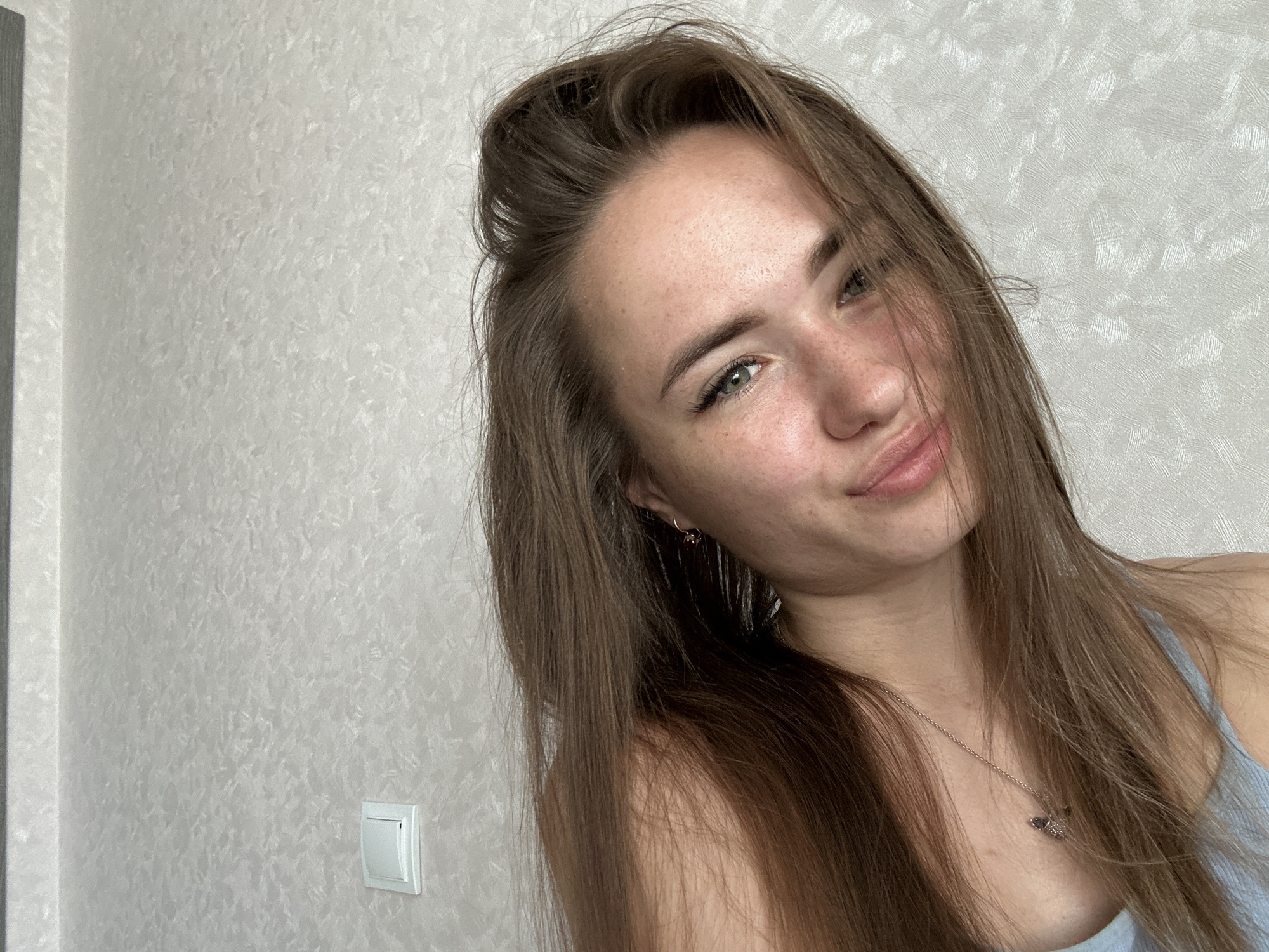 A young woman with long, brown hair and green eyes is pictured from the chest up, looking towards the camera with a slight smirk. She is wearing a light blue top and a silver necklace with a fish-shaped pendant. Her hair is partially covering her face and shoulder. The background is a textured, light-colored wall with a white light switch on the right side. The image appears to be a selfie taken indoors.